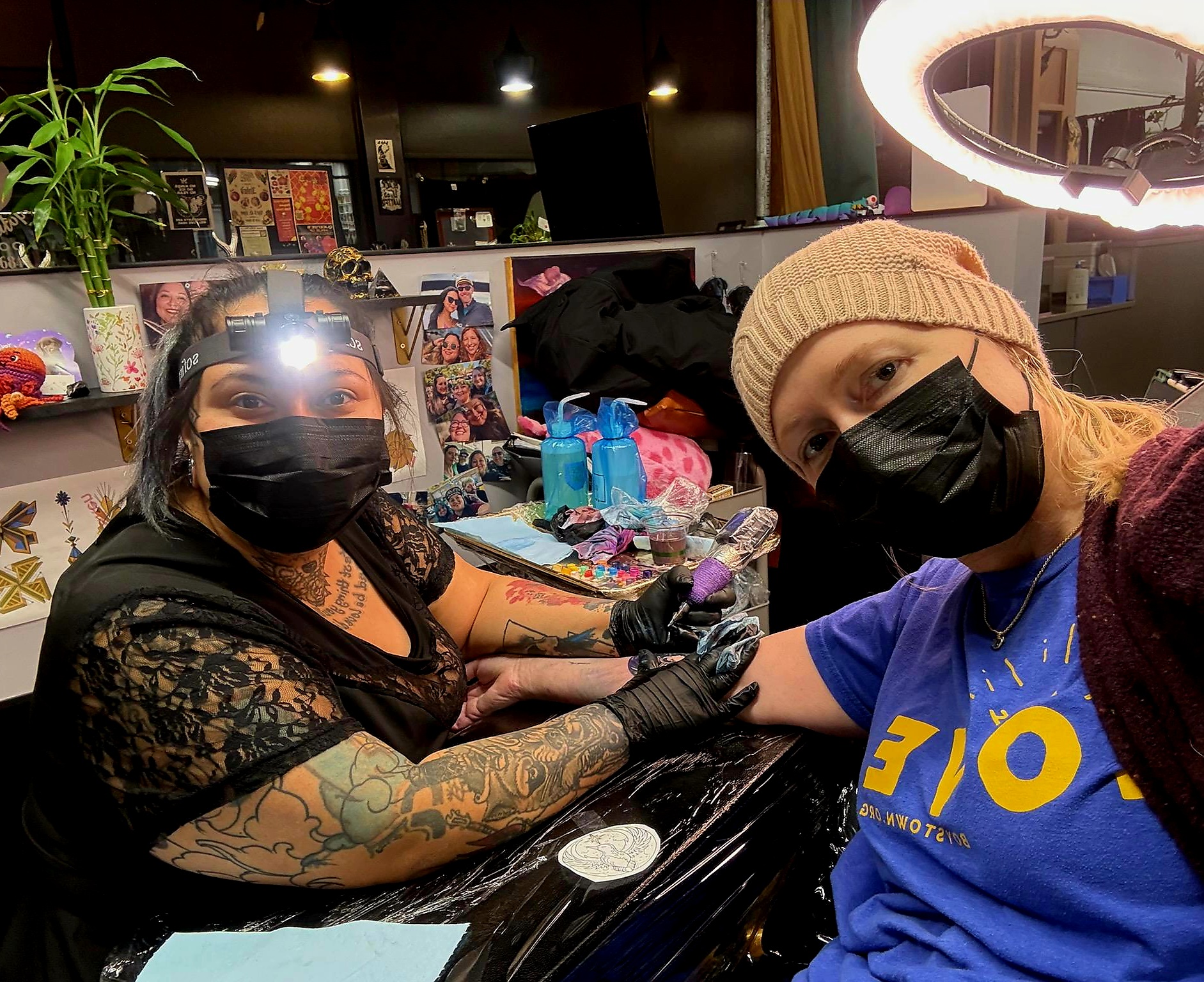 Two women wearing black masks and gloves, one with tattoos, sit at a table with tattoo supplies, in a tattoo studio.