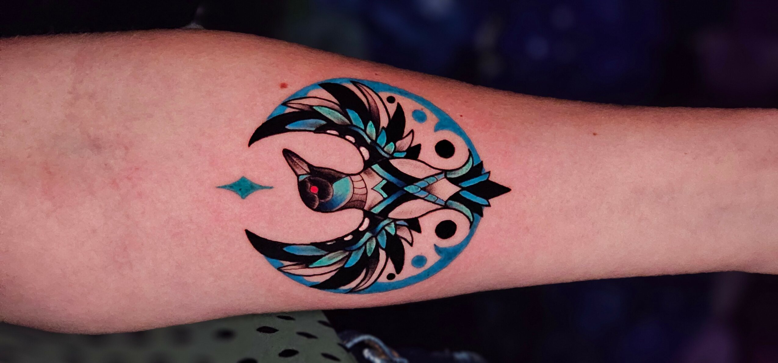 Tattoo of a stylized fish with an arrow, surrounded by abstract shapes and a crescent moon, on a person's arm.