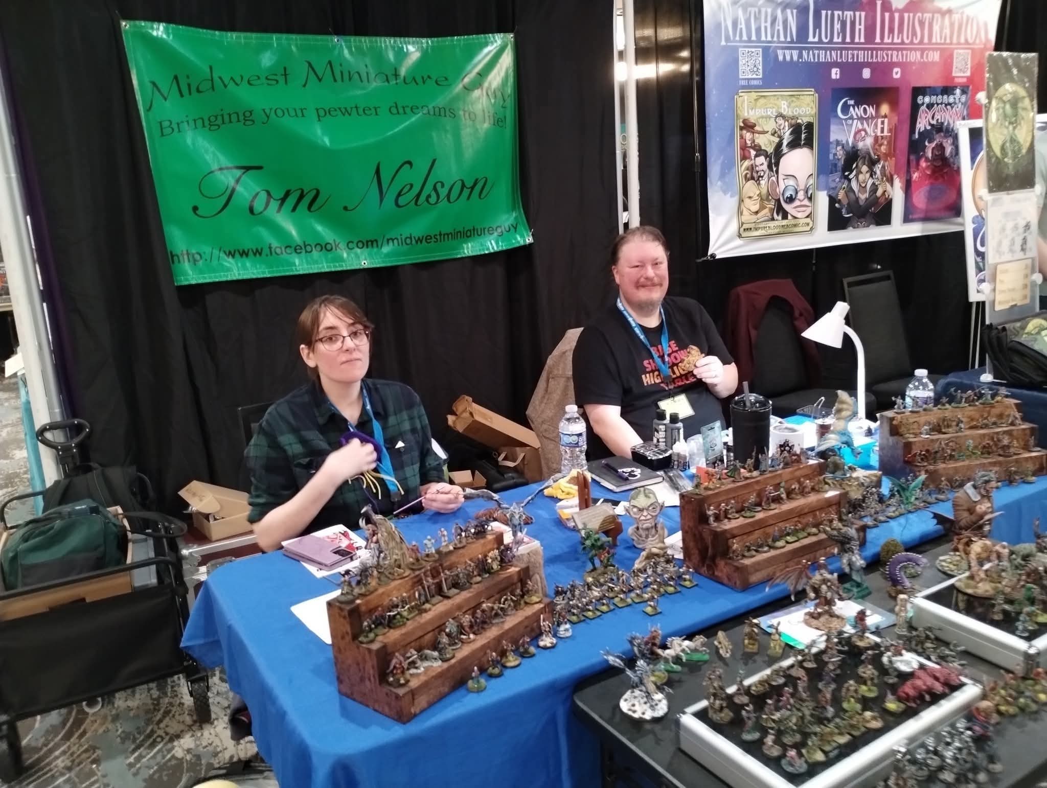 Two people sit behind a table with miniature models, at a booth with banners and posters in the background.