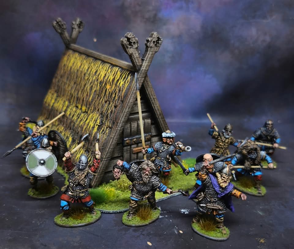 Miniature figures of warriors in armor with weapons, attacking a thatched hut, set on a dark background.
