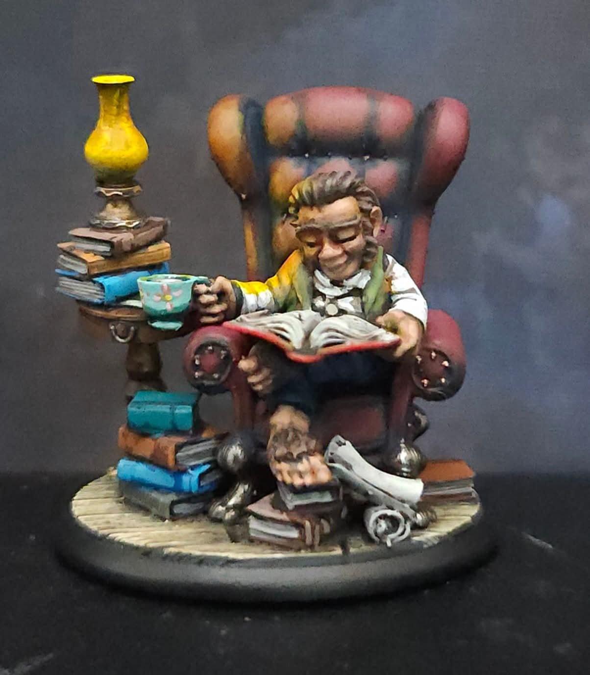 Figurine of a person reading a book, sitting in a large armchair surrounded by stacks of books and a rolled scroll, with a yellow vase on a side table.
