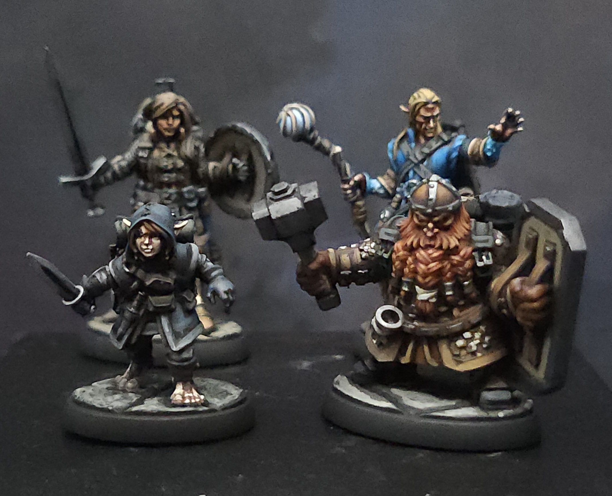 Four painted fantasy miniatures, including a dwarf with a hammer and shield, a wizard with a staff, and two armored figures, on black background.