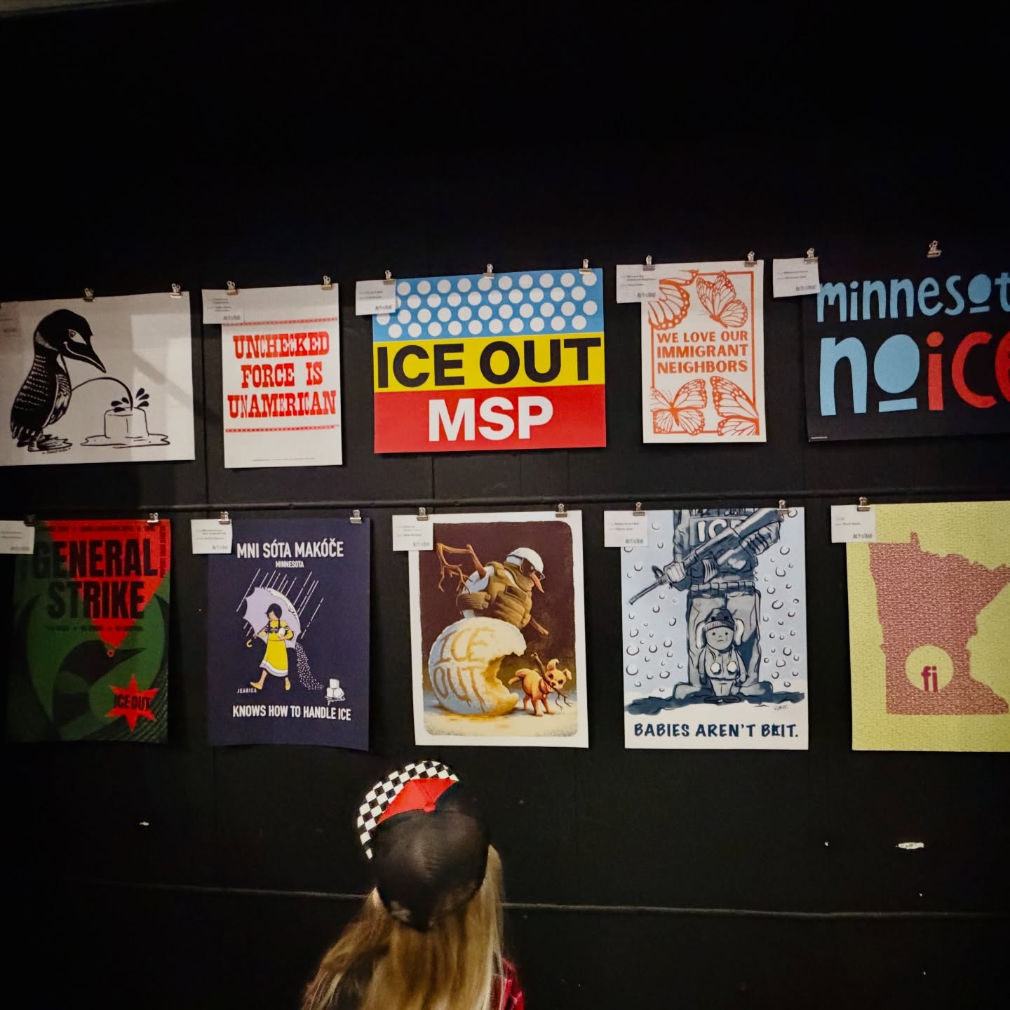Display of various posters on a wall, with a person wearing a checkered hat in the foreground.