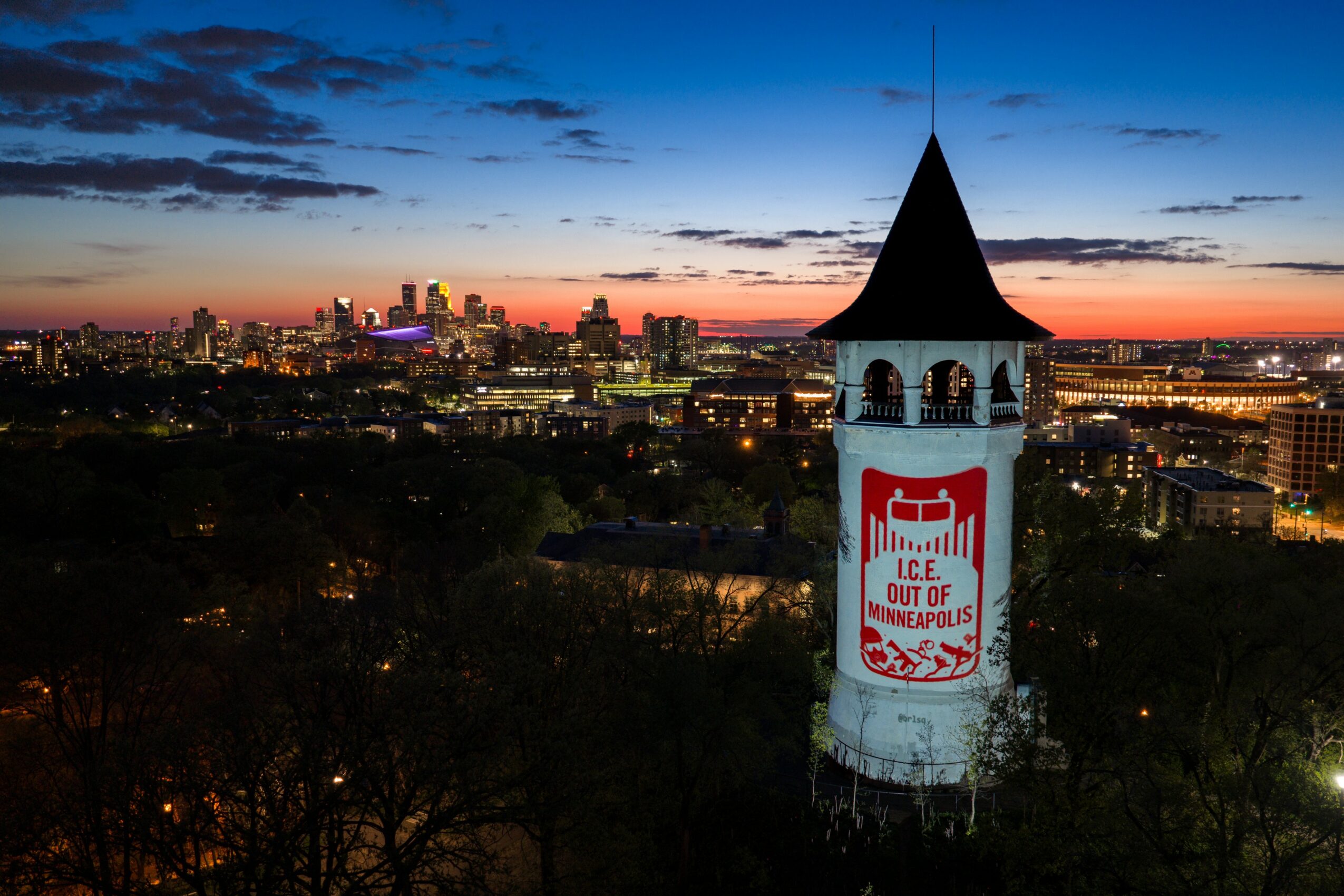 City skyline at sunset with a tall white tower displaying a red sign that says 'ICE OUT OF COMPLAINY' and trees in foreground.