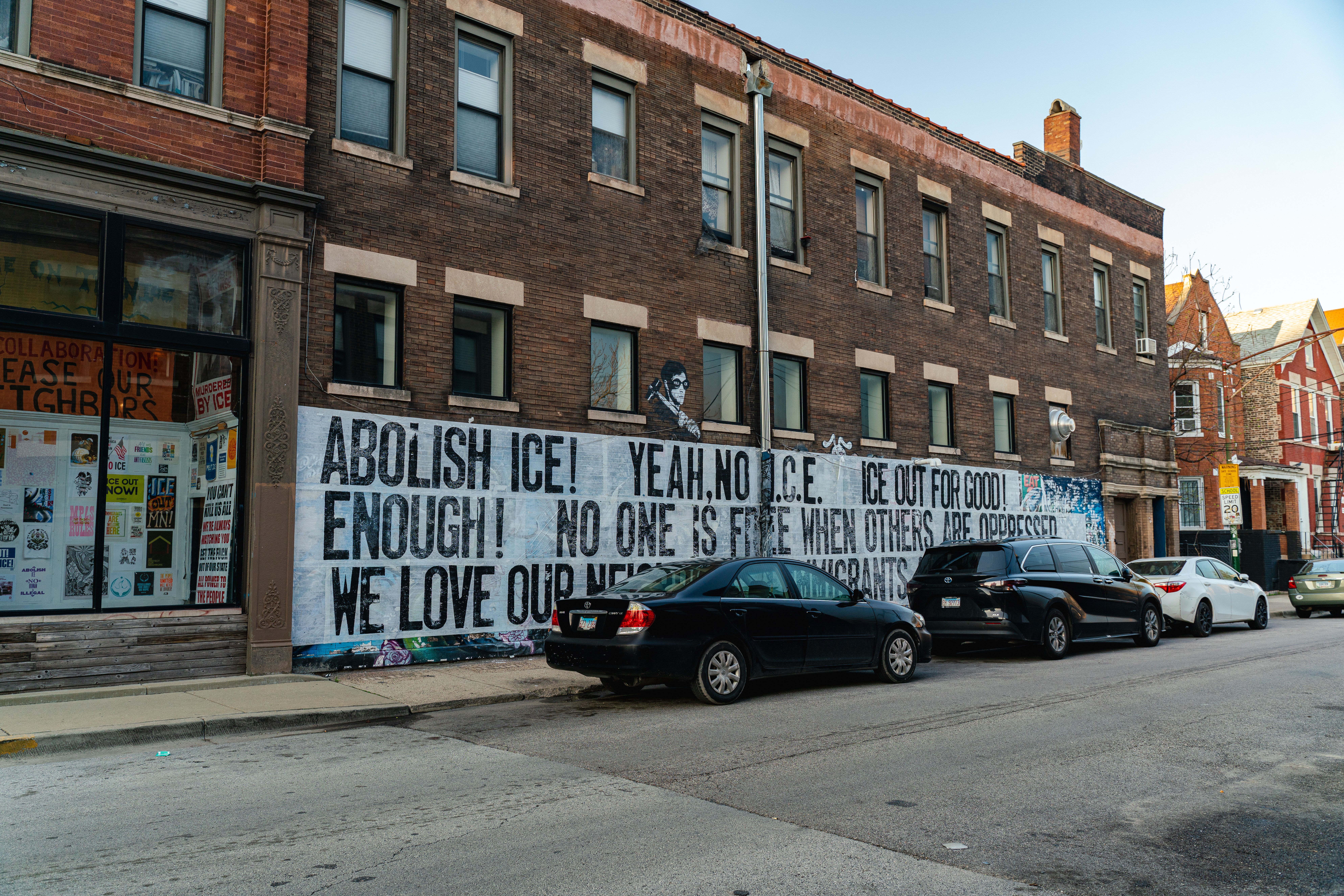 Brick building with a large banner protesting ice, with parked cars in front, on a city street.