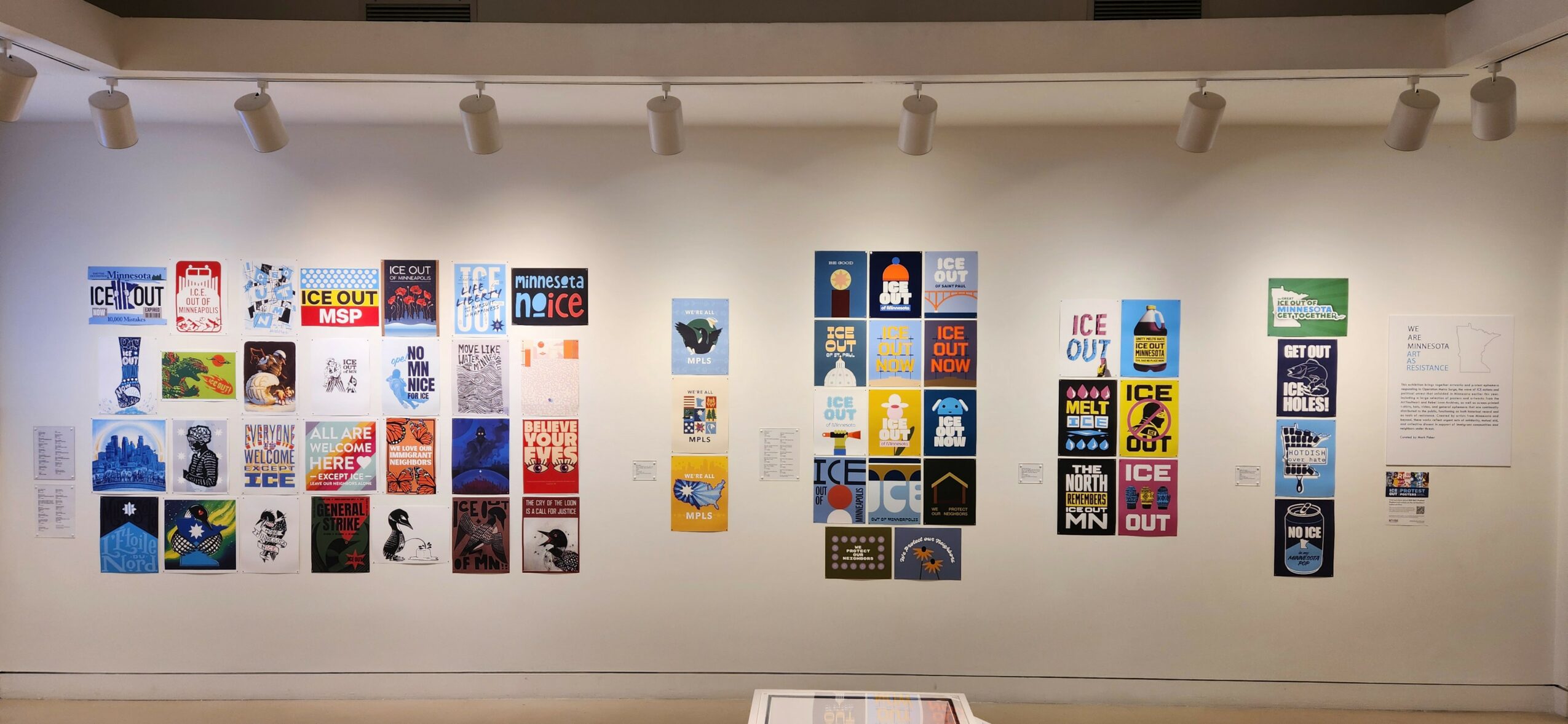 Gallery wall with colorful posters and artwork arranged in rows, displayed on a white wall with ceiling lights.