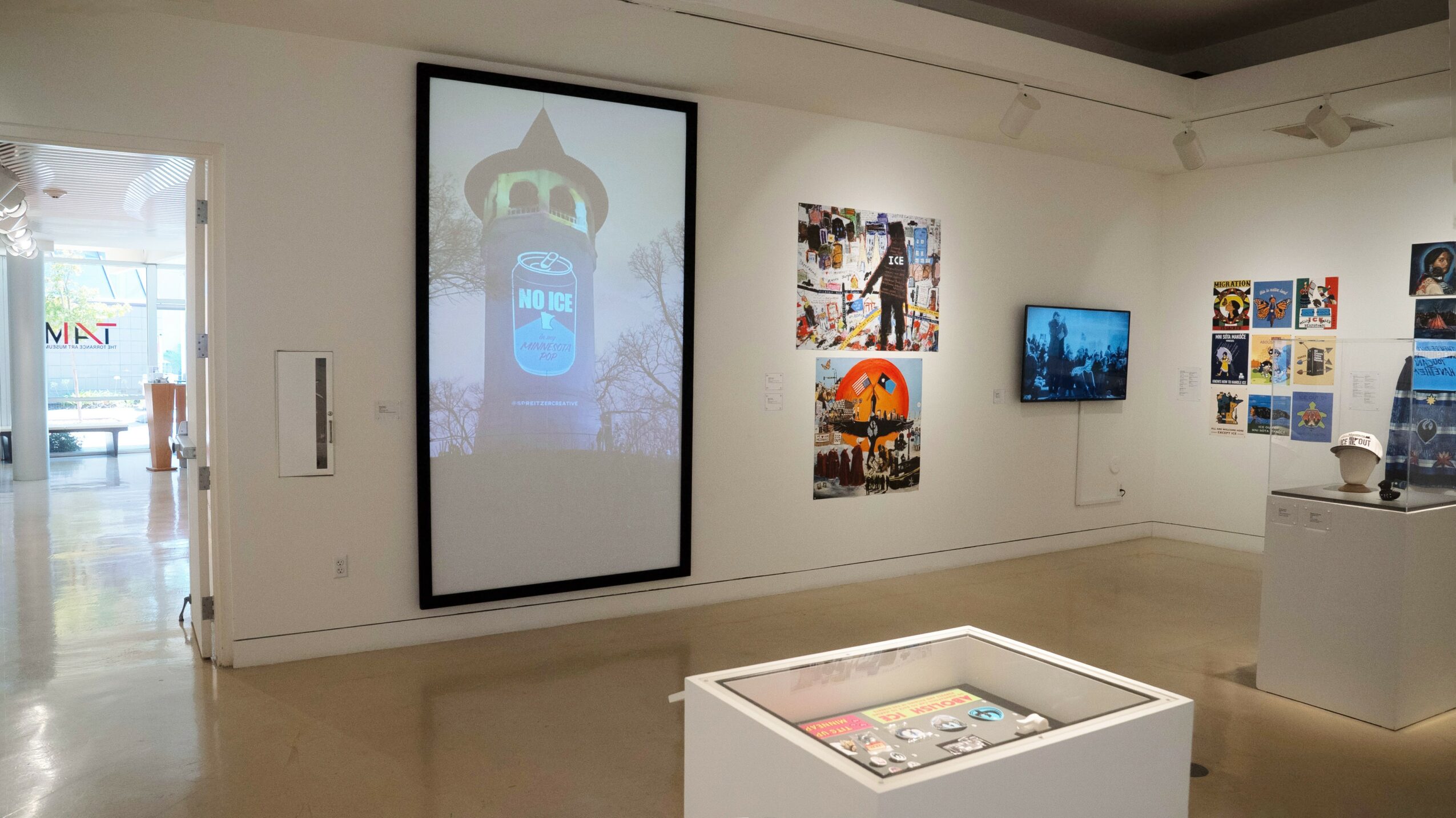 Gallery with digital screens and artwork on white walls, a glass display case in the center, and a doorway to another room.