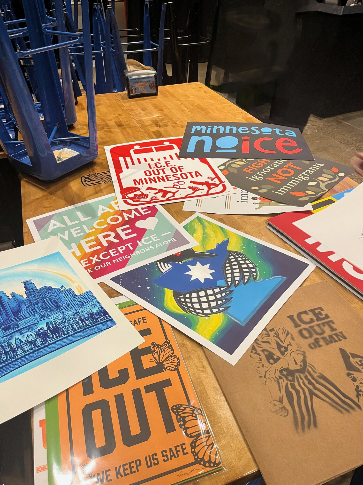 Various colorful postcards and flyers spread on a wooden table, some with text and images, with blue chairs in the background.