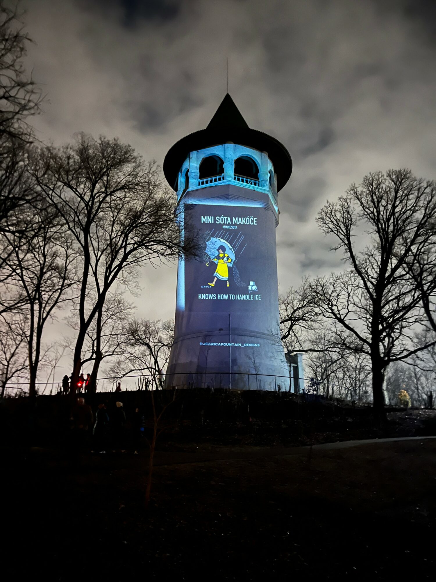 Tall tower with a pointed roof illuminated at night, surrounded by leafless trees and cloudy sky.