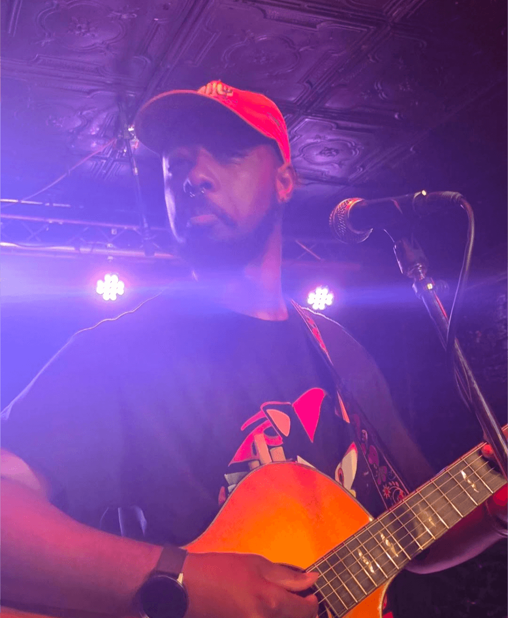 Person wearing a red cap playing an acoustic guitar on stage with purple lighting, microphone nearby.