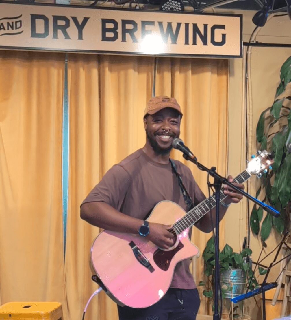 Man playing guitar and singing into microphone on stage with yellow curtains and a sign that reads 'DRY BREWING'.