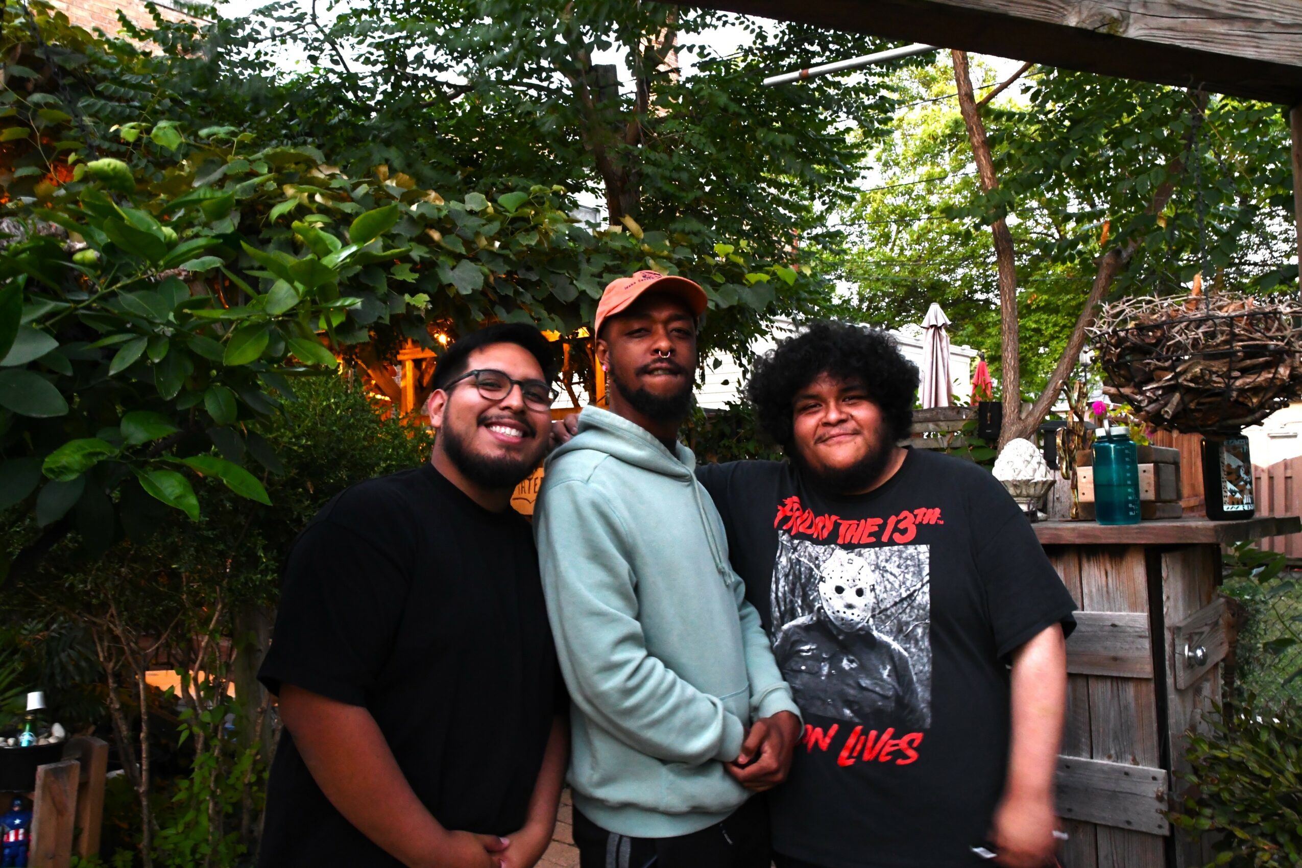 Three people standing outdoors in a garden, smiling, with trees and a wooden fence in the background.