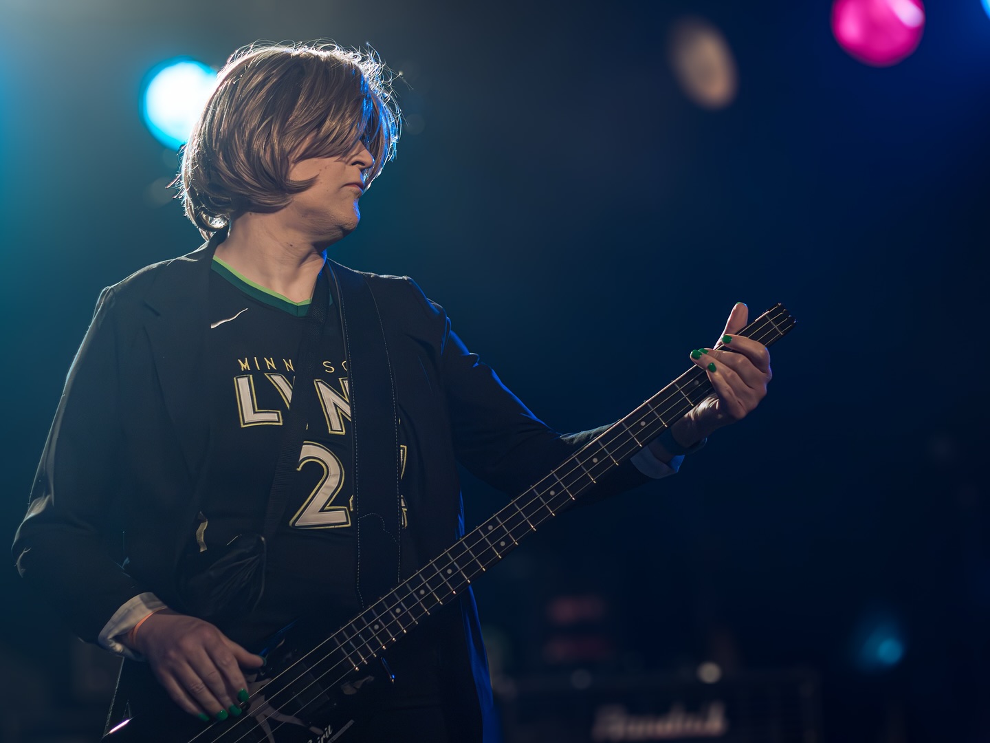 Person with shoulder-length hair playing an electric guitar on stage with colorful lights in background.
