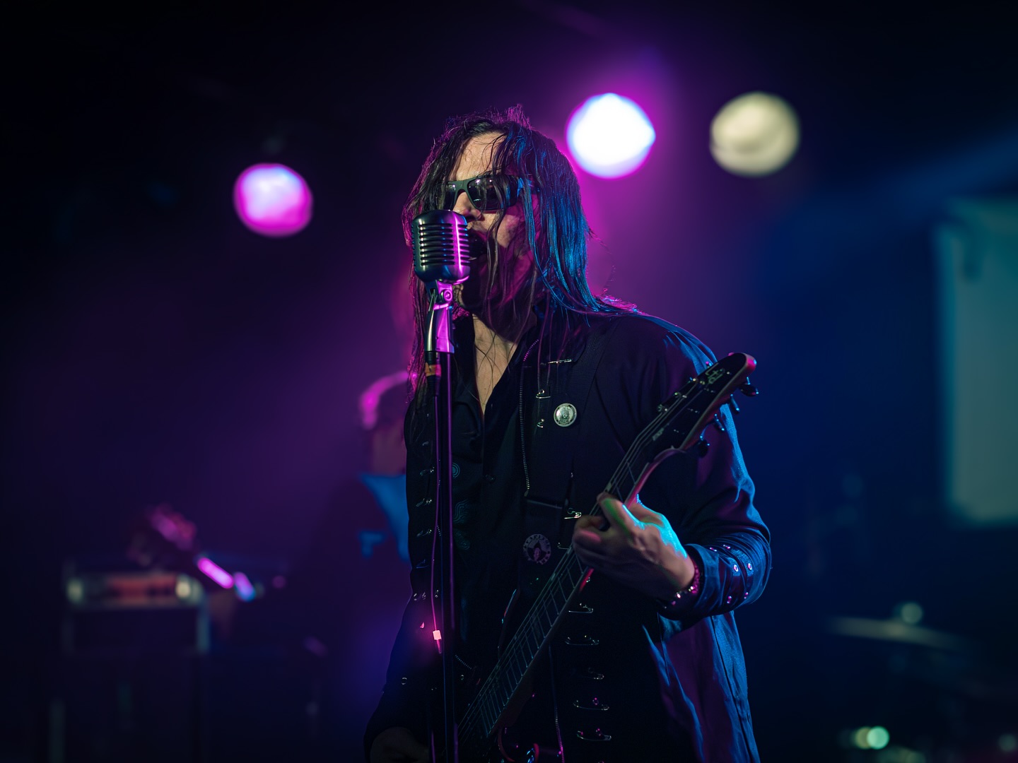 Person with long hair and sunglasses singing into a vintage microphone while playing guitar on stage with colorful lights