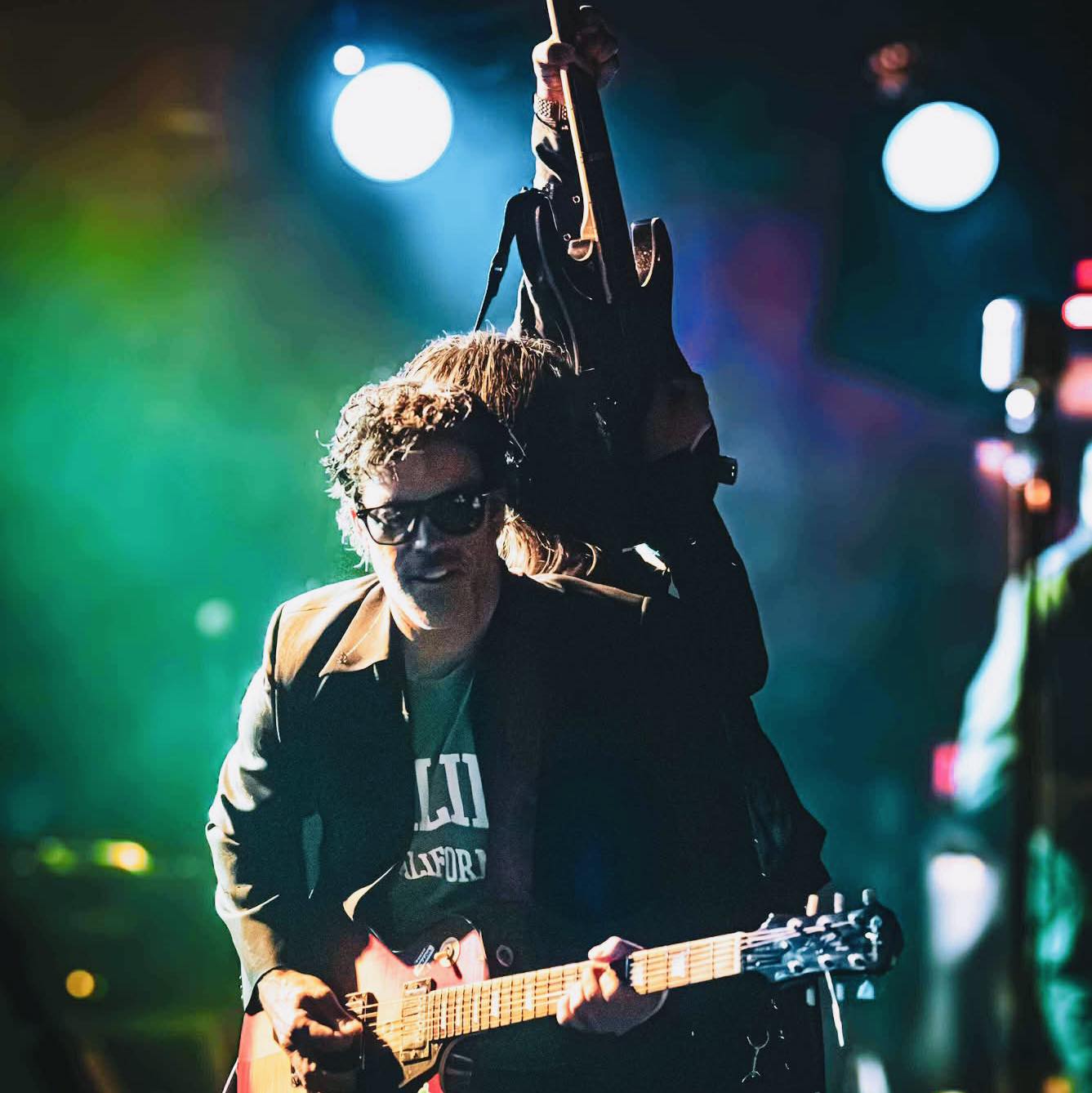Person playing guitar on stage with colorful lights and a dark background.