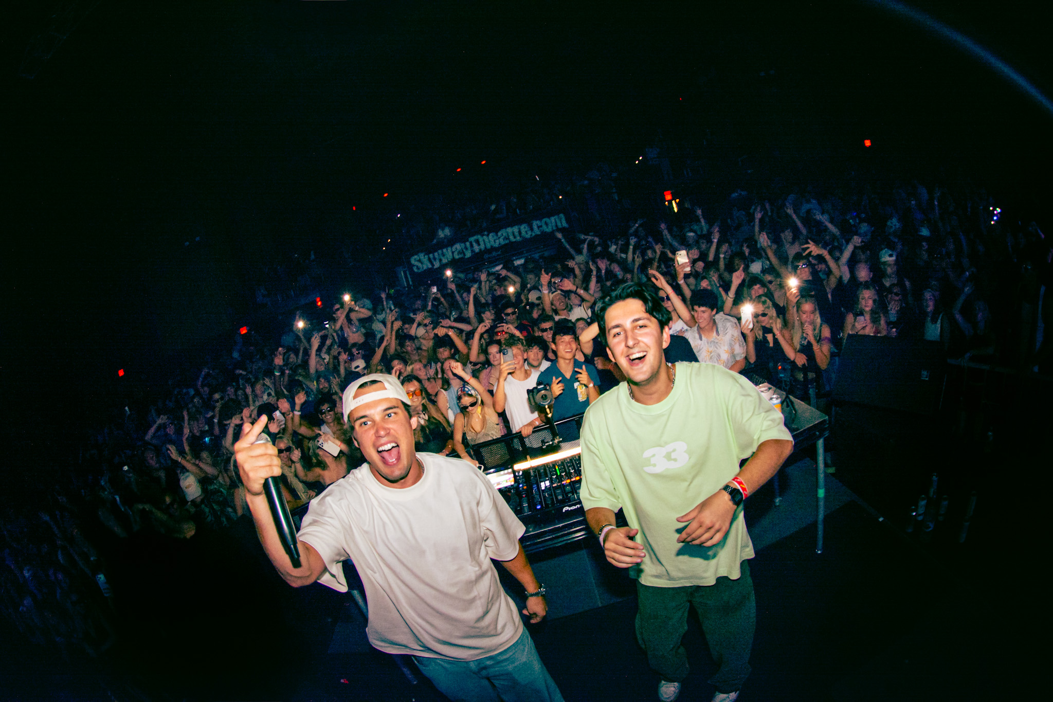 Two young men smiling and waving at a crowded concert or party with many people and lights in the background.