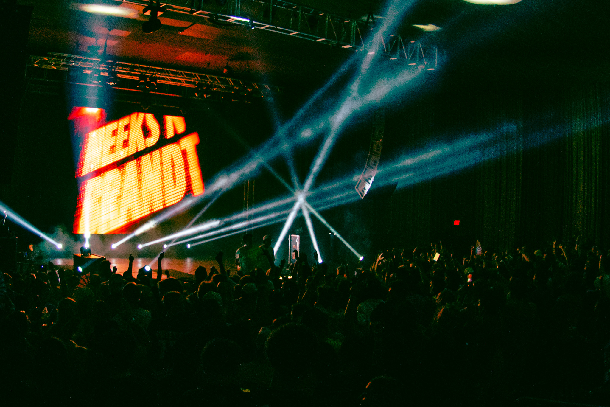 Concert stage with bright lights and a large screen displaying 'TEENS IN TANDT', audience in foreground, dark atmosphere.