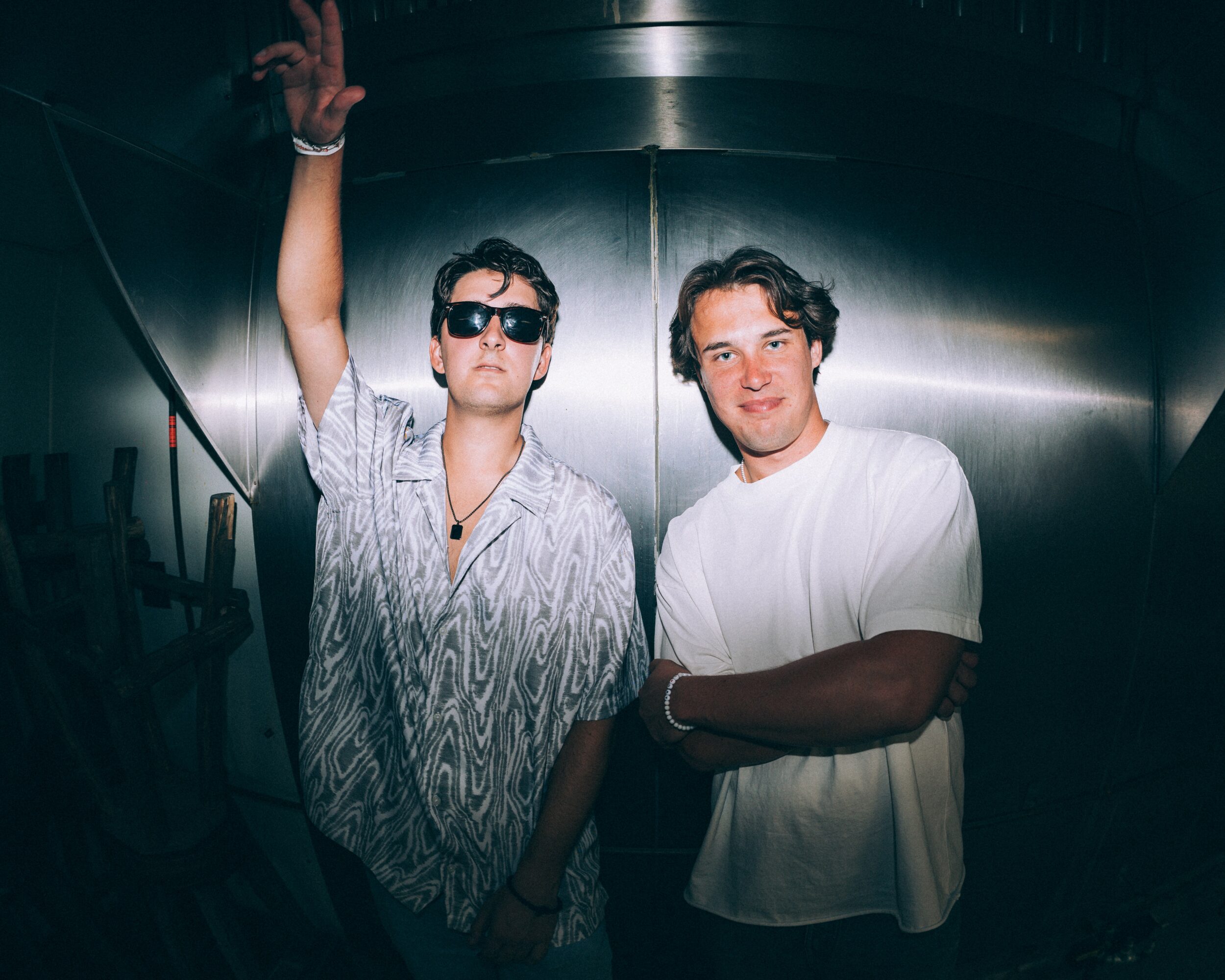 Two men stand in a metallic room, one wearing sunglasses and a patterned shirt, the other in a white shirt with arms crossed.
