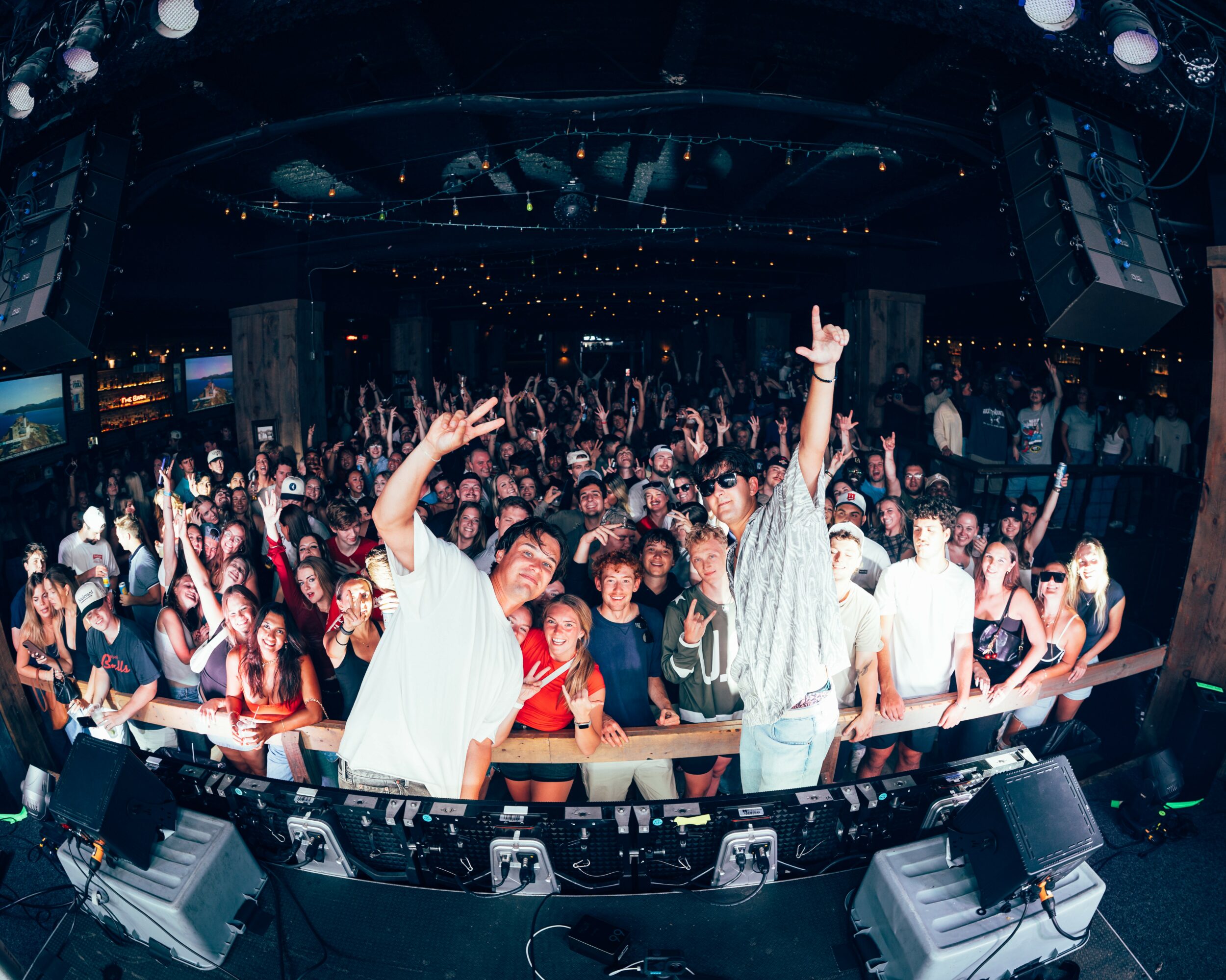 Crowd of people at a concert or club, with two performers on stage in front, one raising an arm, others dancing.
