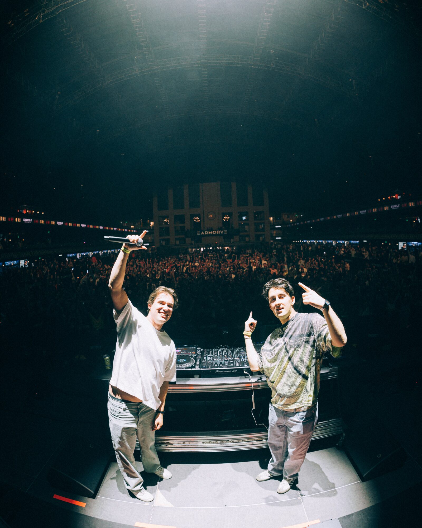 Two people standing behind a DJ booth in a large indoor venue, with a crowd and stage in the background.