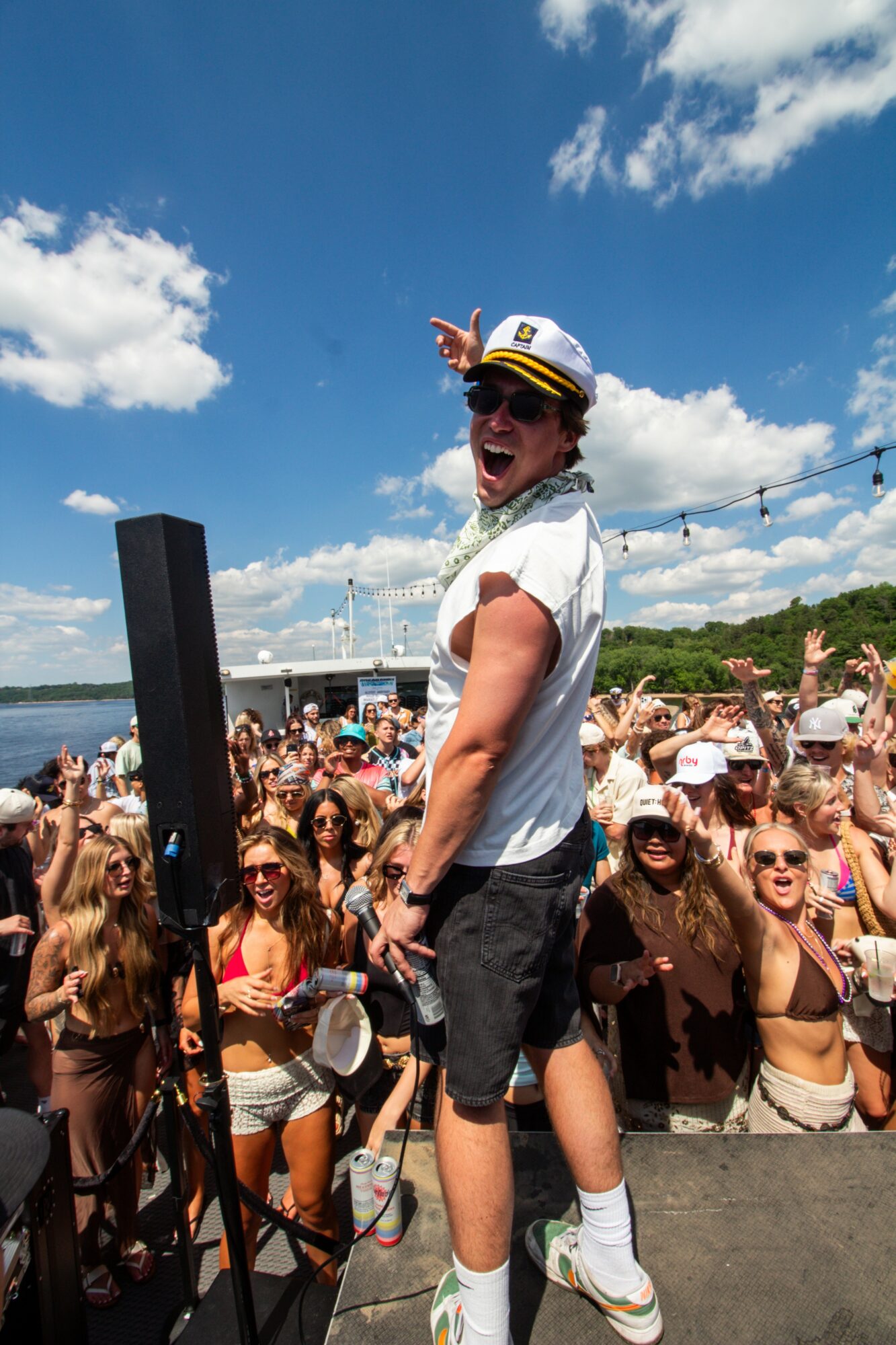Person wearing a white shirt, shorts, and a cap, smiling and making a peace sign at an outdoor event with a crowd, sky, and water in background.