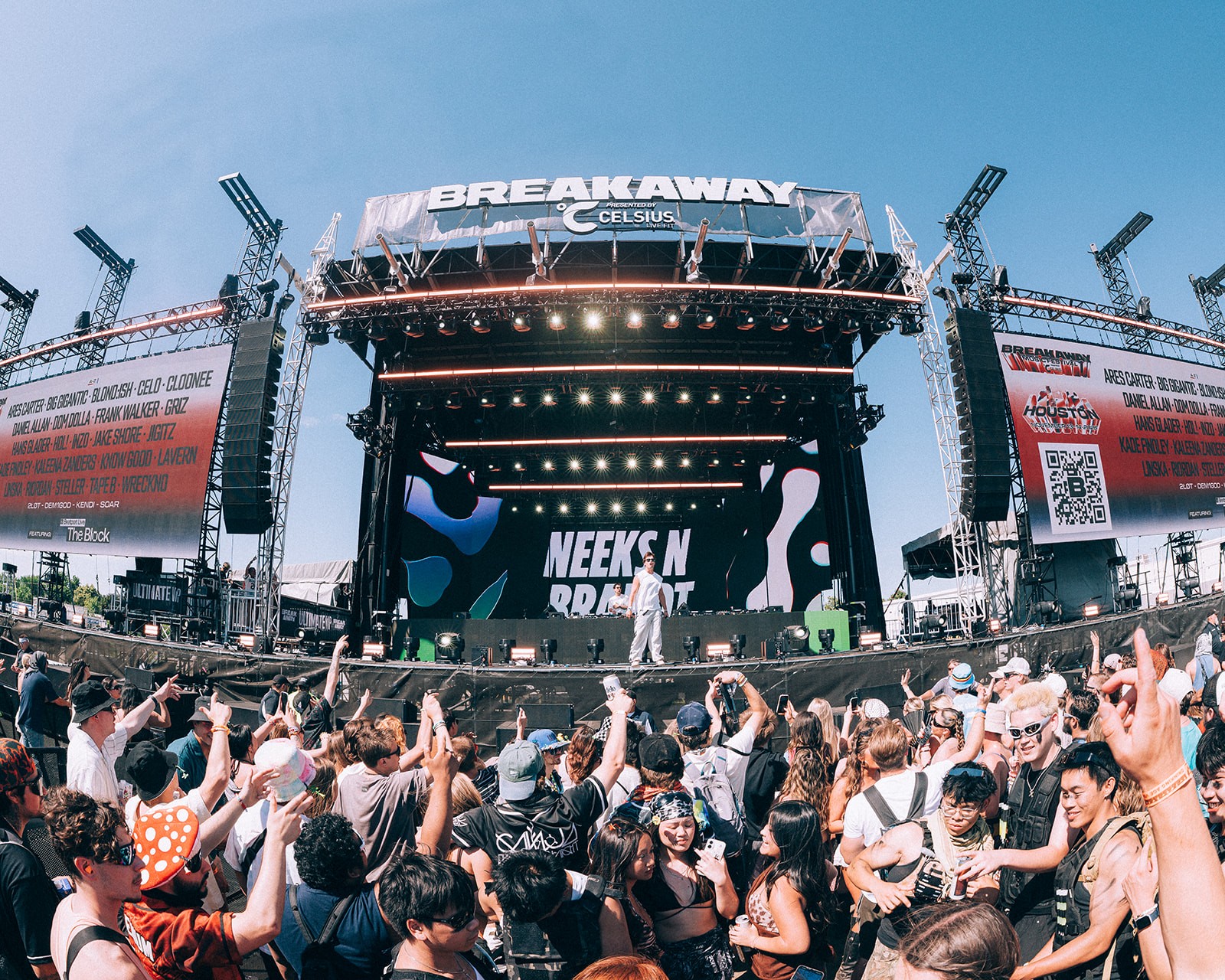 Large outdoor concert stage with crowd, stage lights, and screens, under a clear blue sky.