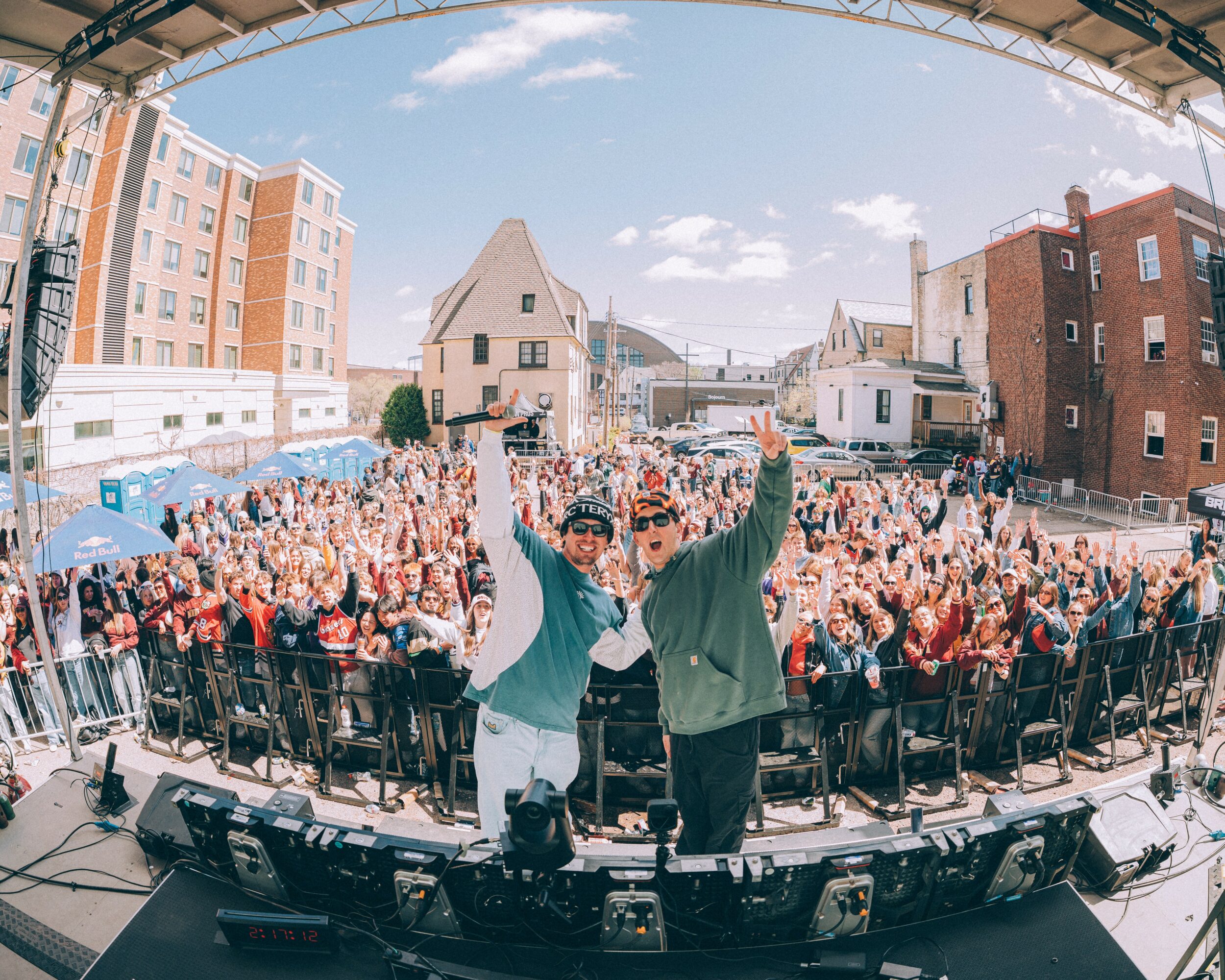 Two performers on stage with a large crowd outdoors, buildings in the background, under a partly cloudy sky.