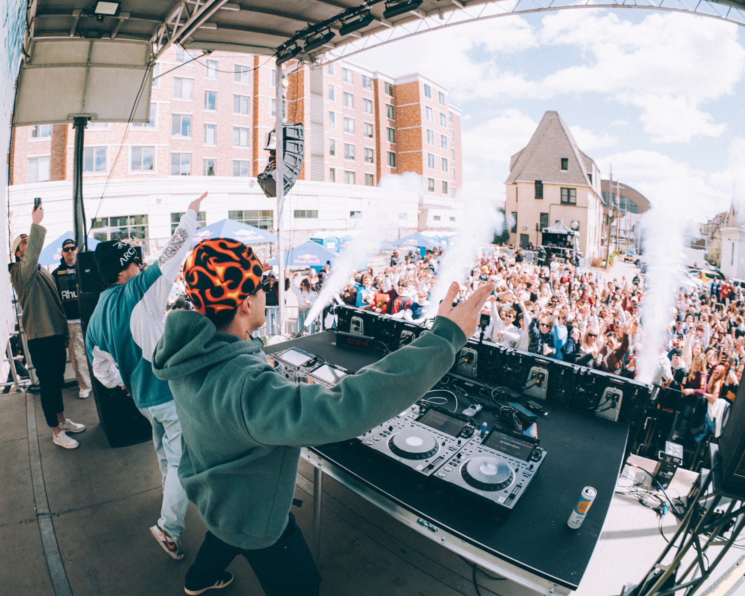 DJ performs on stage with a crowd outdoors, buildings in the background, and smoke effects, under a large arch.