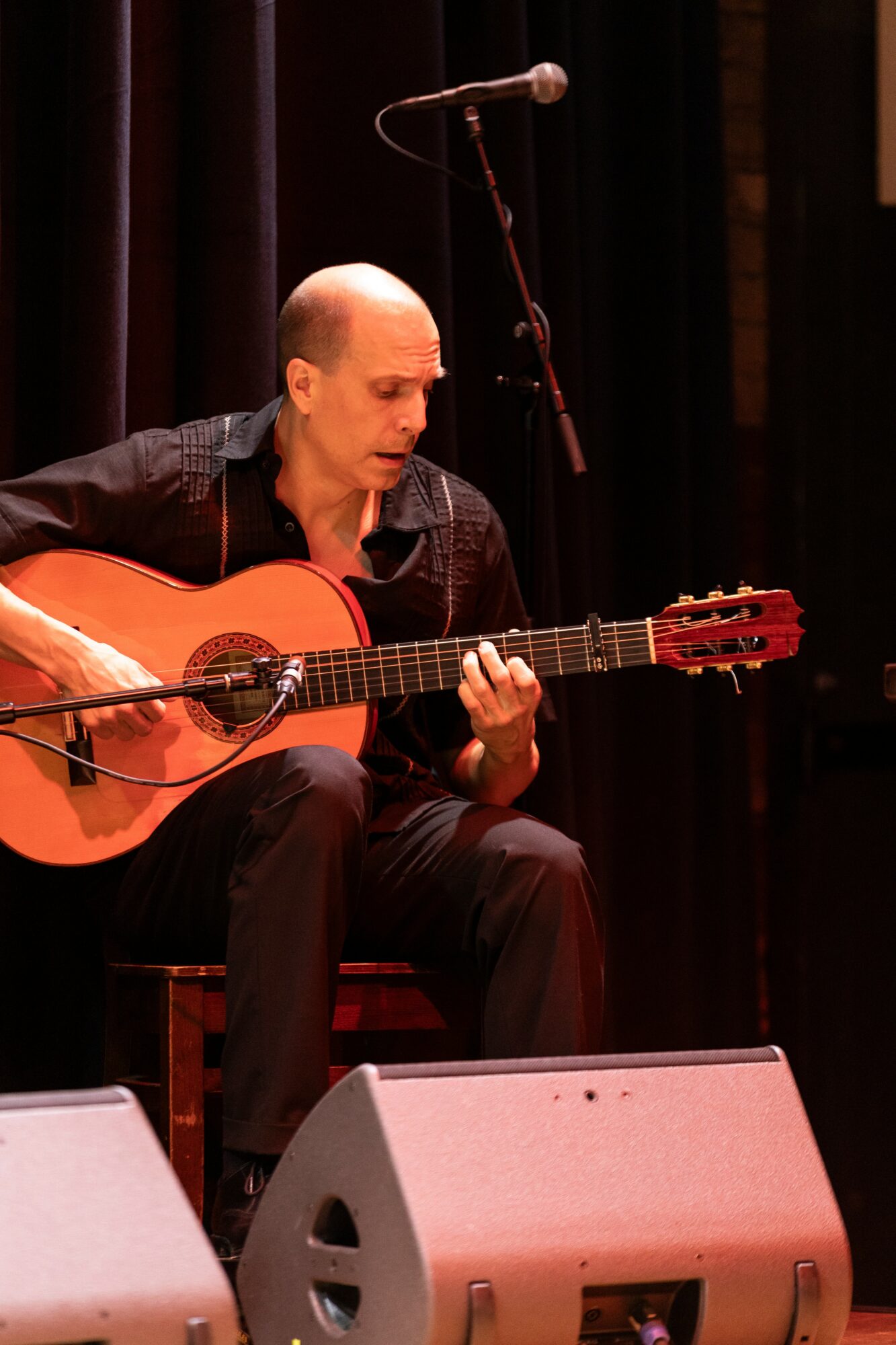 Bald man playing an acoustic guitar on stage with a microphone overhead, dark background, stage monitors in front.