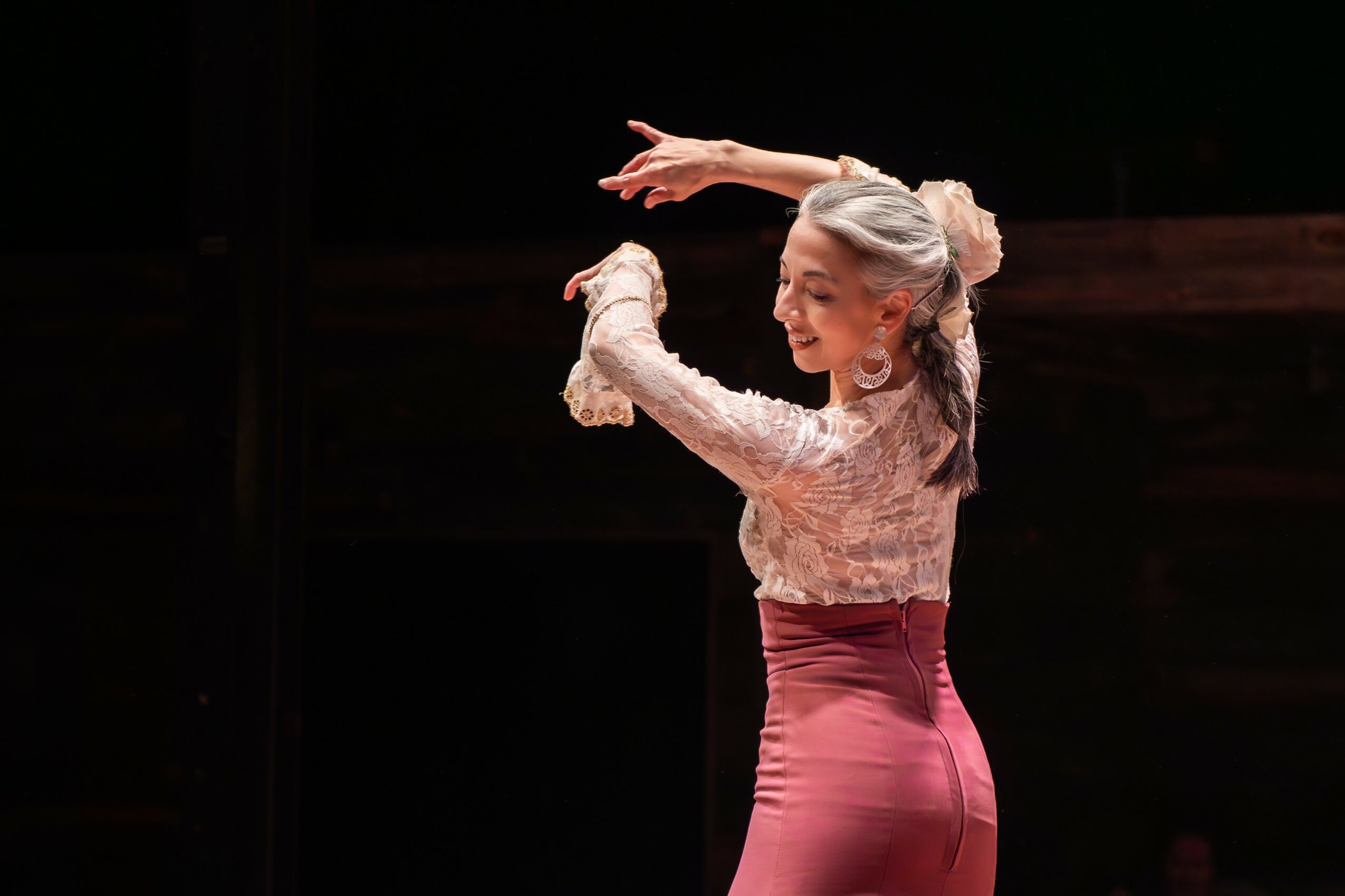 Young girl with gray hair in a bun, wearing a lace top and pink skirt, dancing on stage with arms raised.