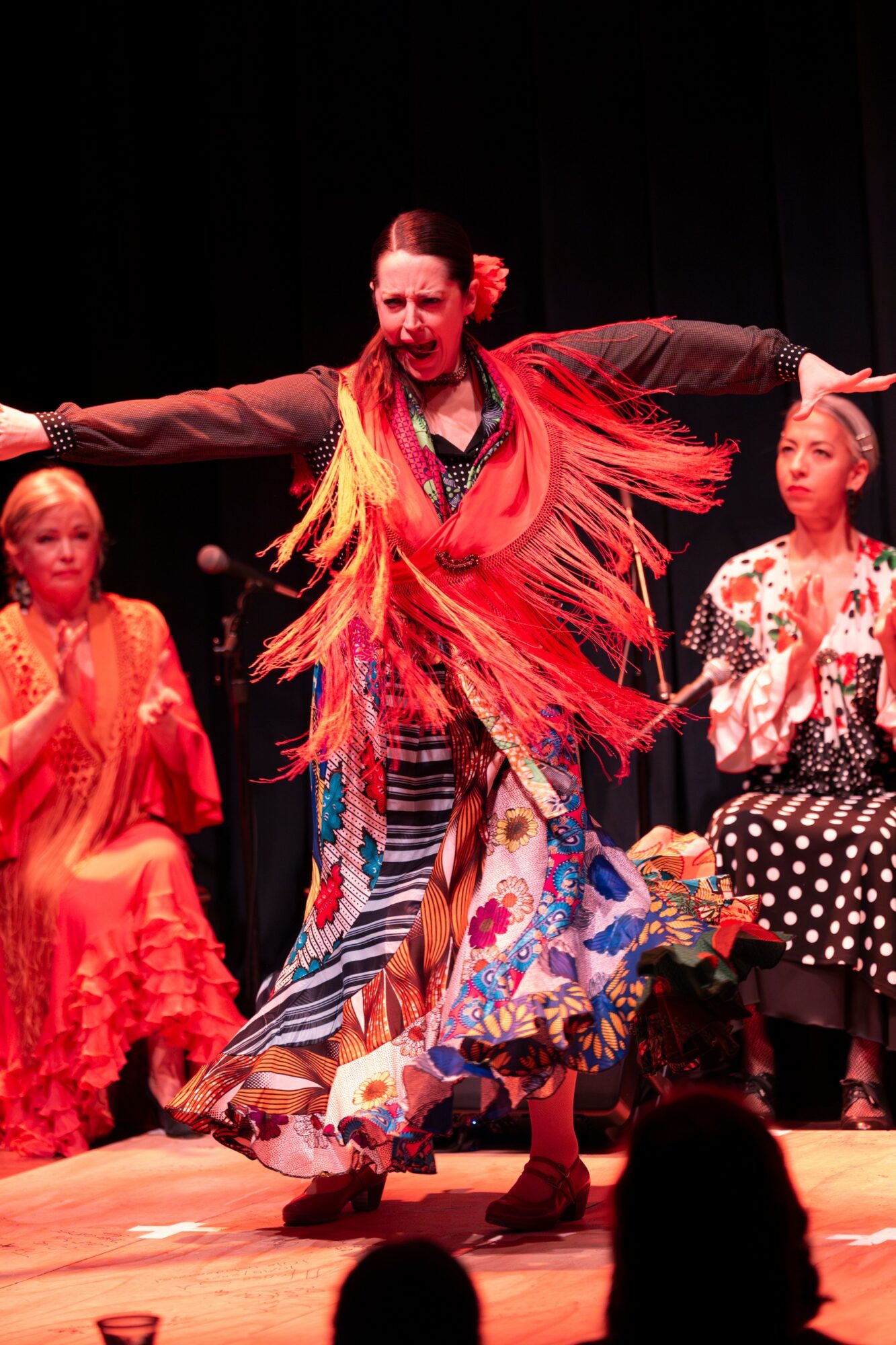 Performer in colorful costume dancing on stage with two seated women in traditional attire, dark background, stage lighting.