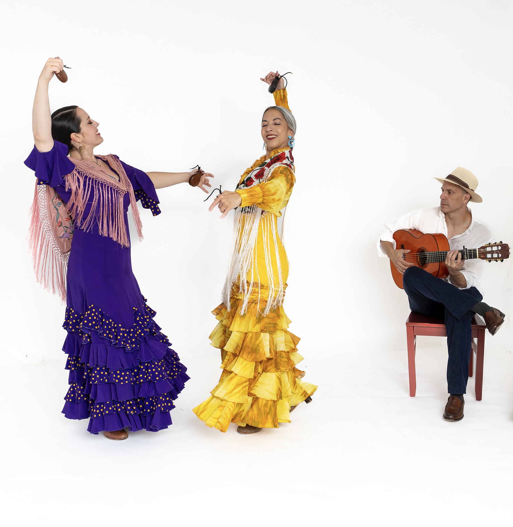 Two women dancing in colorful dresses and a man playing guitar on a red chair, all against a white background.