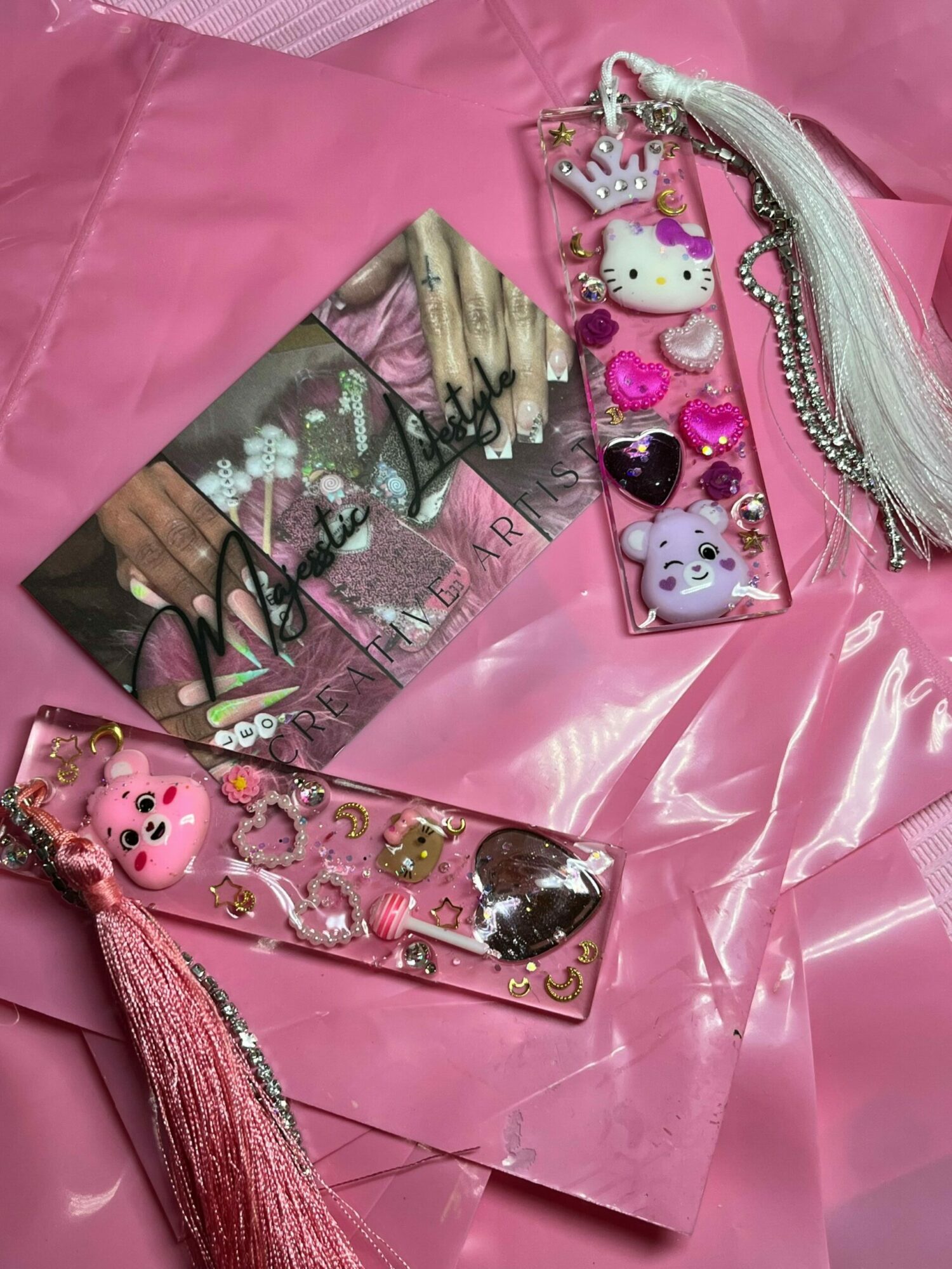 Four decorated phone cases with charms and accessories on pink fabric, featuring Hello Kitty, hearts, and a pink bear, with tassels.