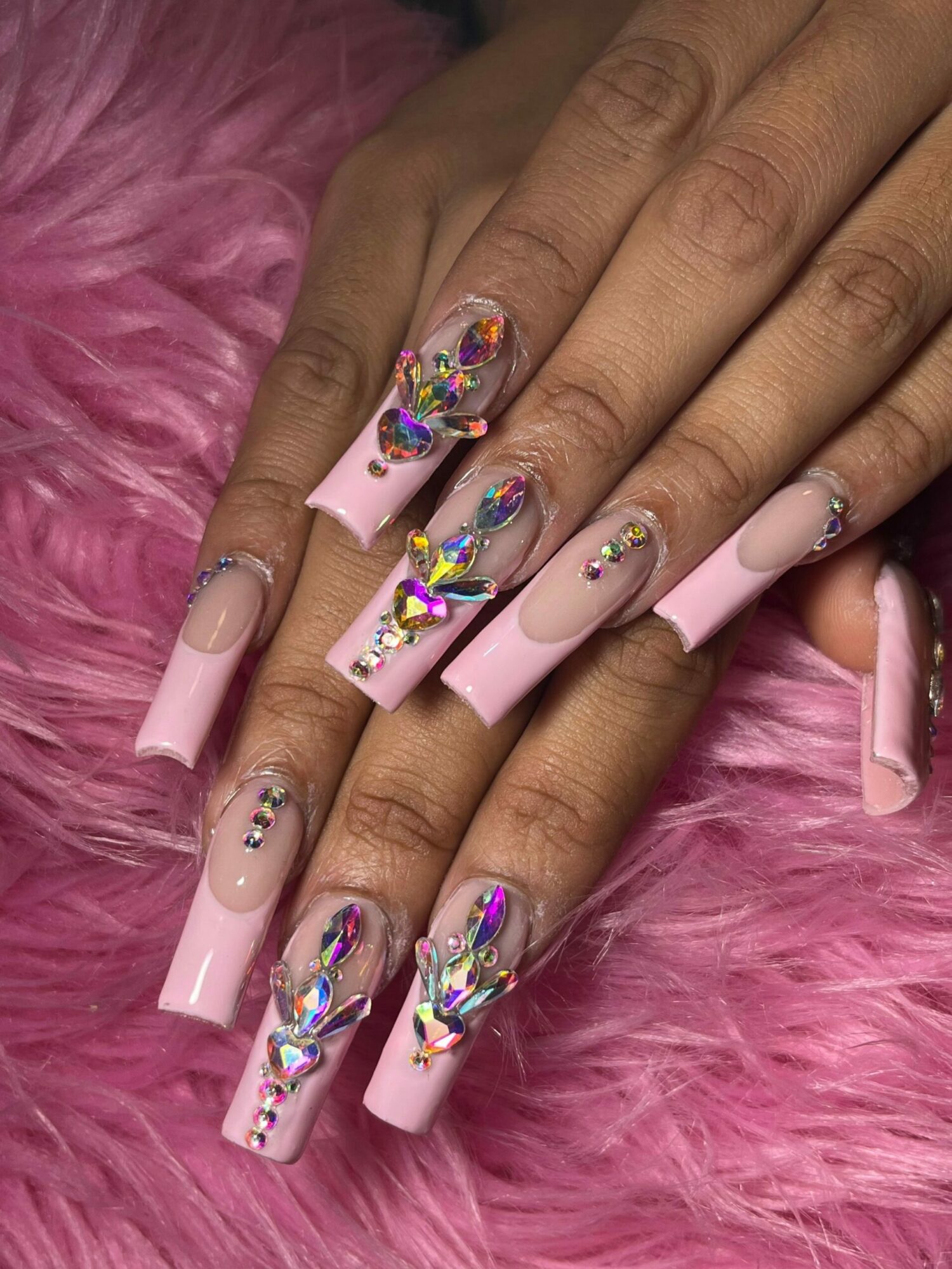 Hands with long, pink, decorated nails resting on pink furry surface, featuring rhinestones and colorful designs.