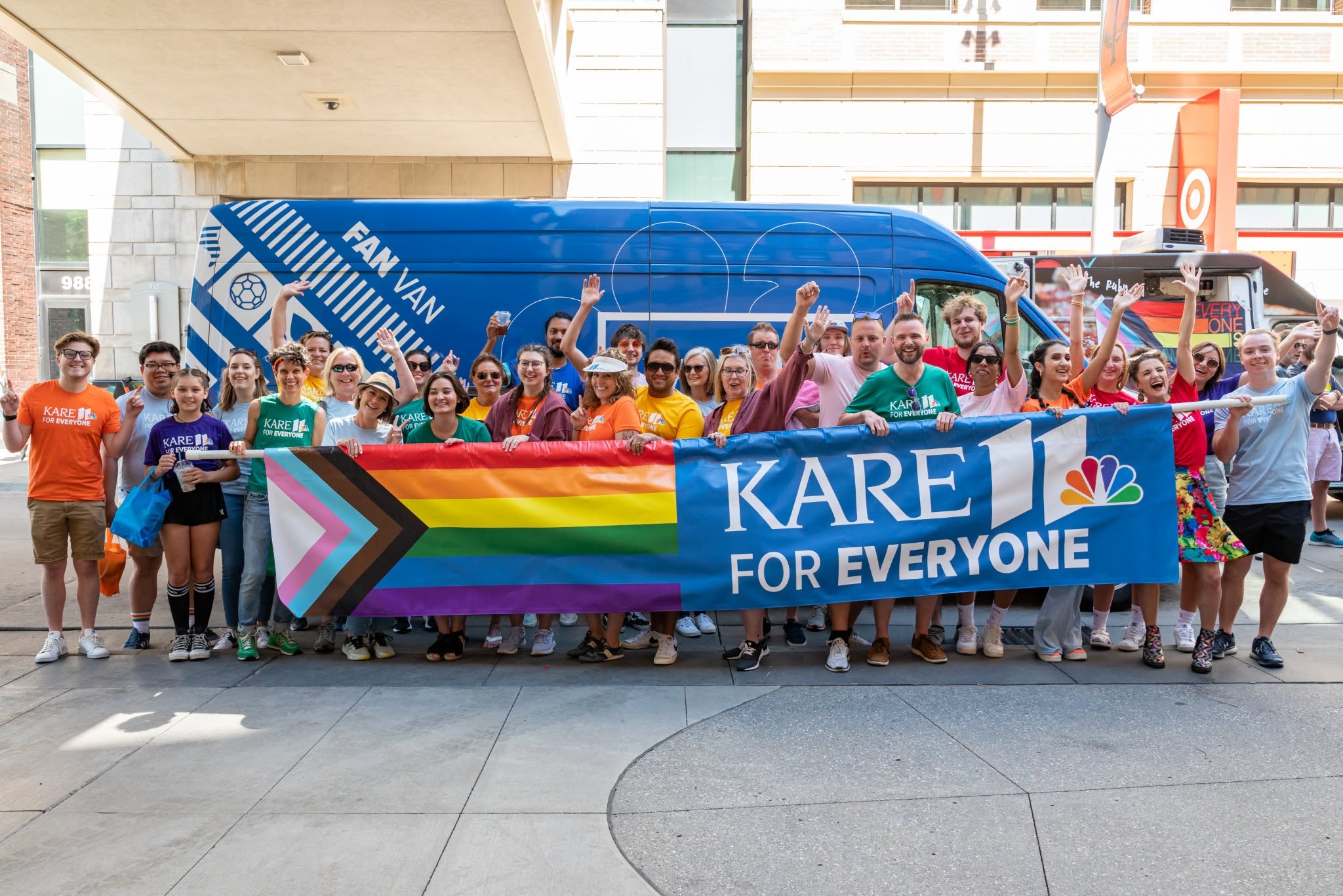 Group of people holding a rainbow flag and a banner that reads 'KARE FOR EVERYONE' in front of a blue bus, outdoors.