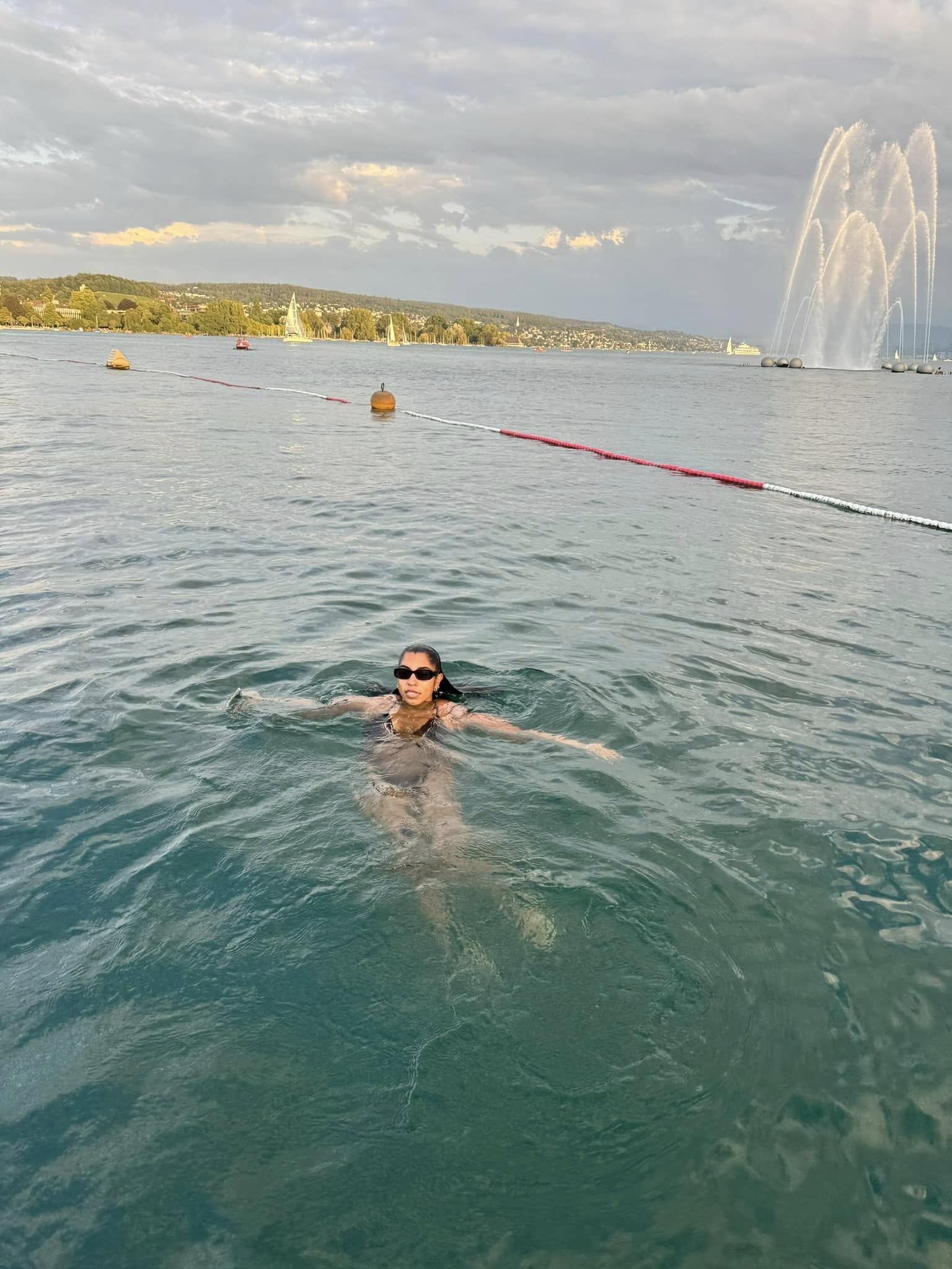 Person swimming in a large body of water with a fountain and distant shoreline in the background.