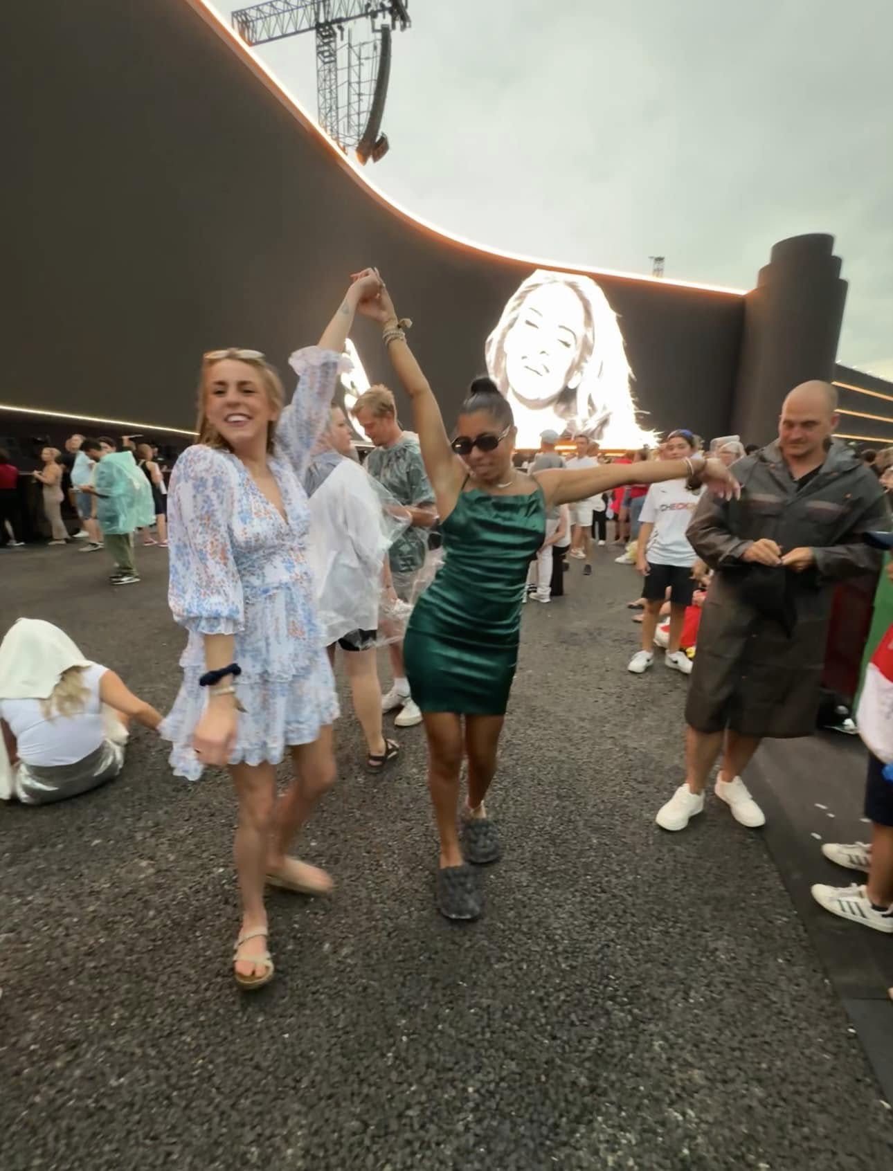 People dancing outdoors with a large screen in the background, some holding hands, smiling, and enjoying the event.
