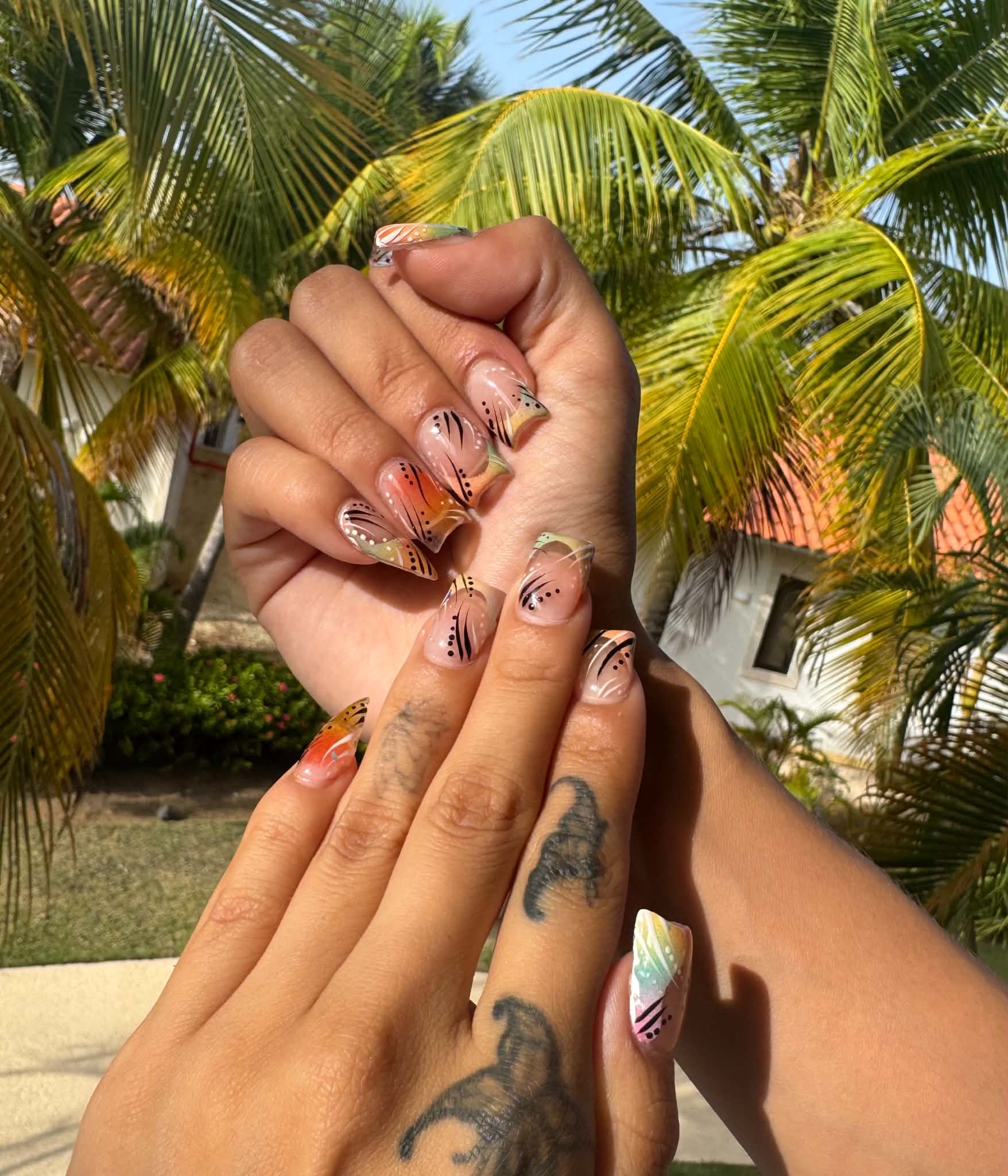 Close-up of hands with colorful nail art, palm trees and a building in the background, sunny outdoor setting.
