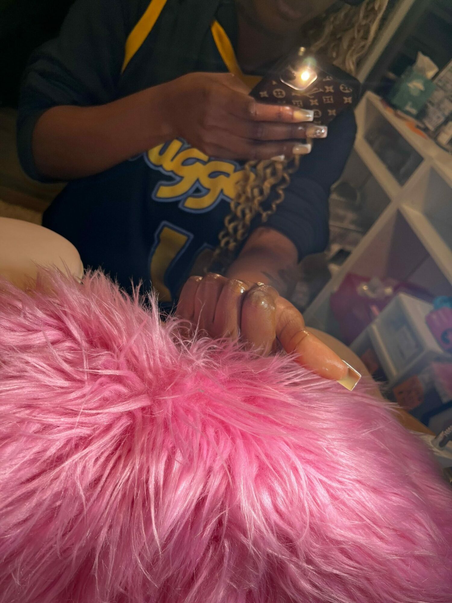Person with pink hair giving a thumbs-up, holding a phone, in a room with shelves and storage bins.