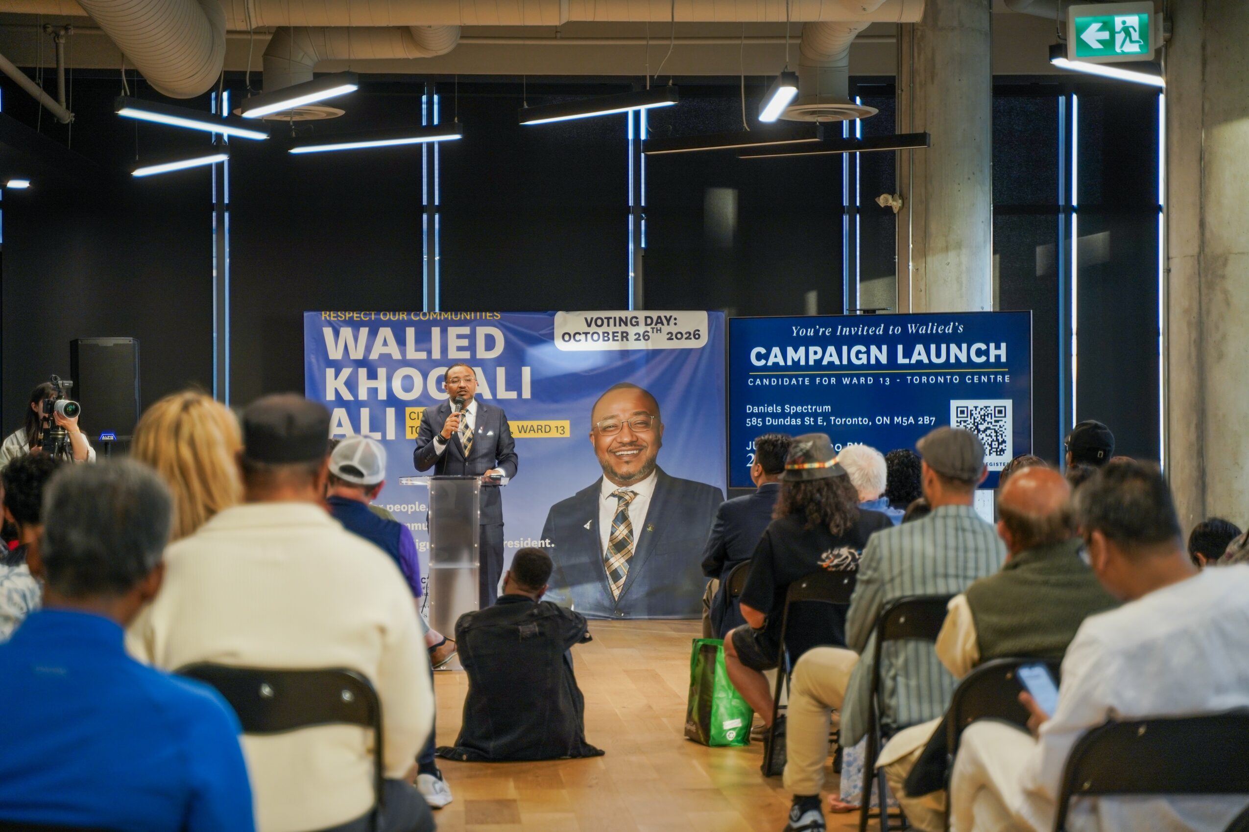 Person speaking on stage at campaign launch event with audience seated, large banner in background, modern interior setting.
