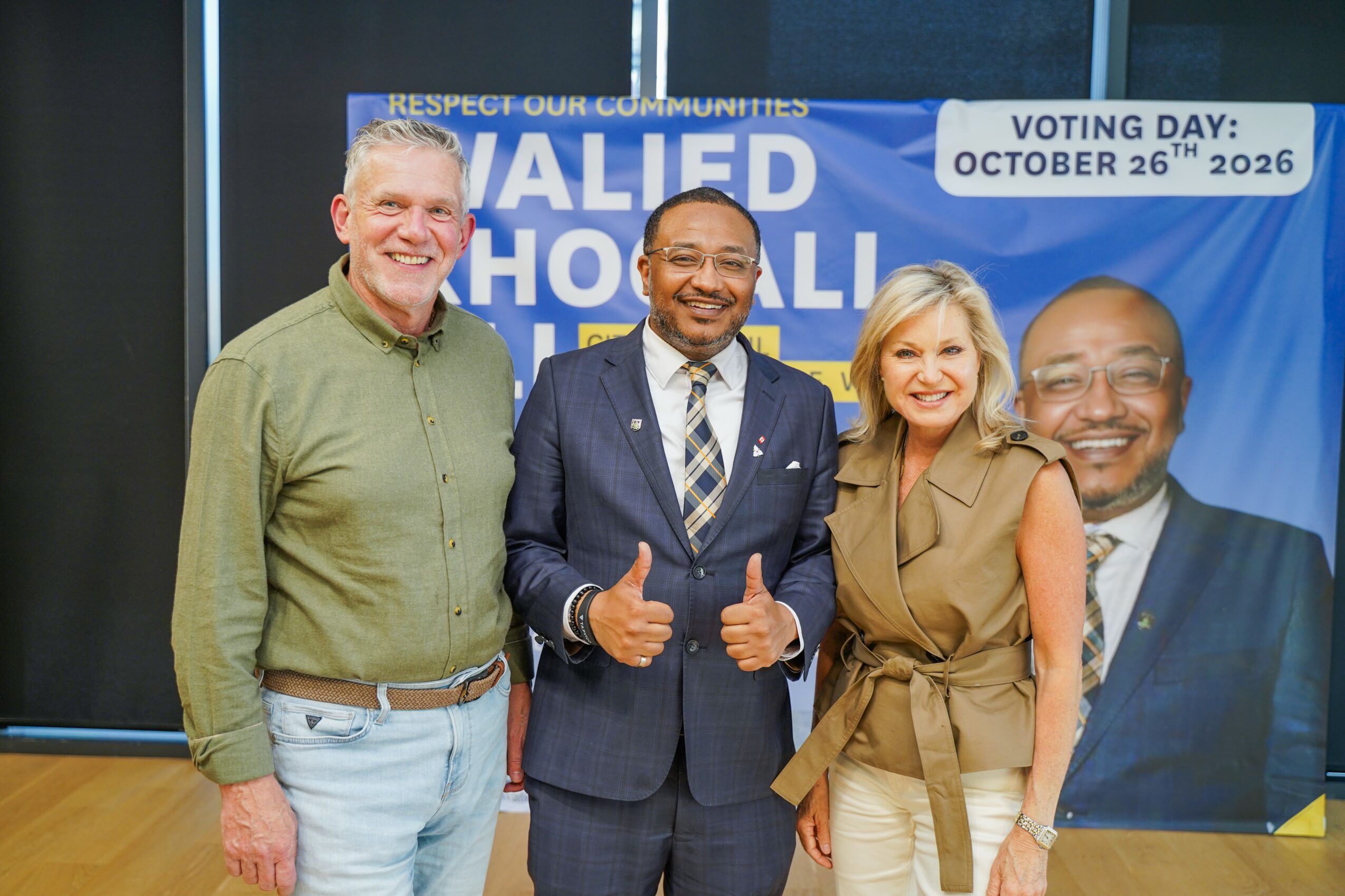 Four people standing together smiling, with a large campaign poster in the background, in an indoor setting.