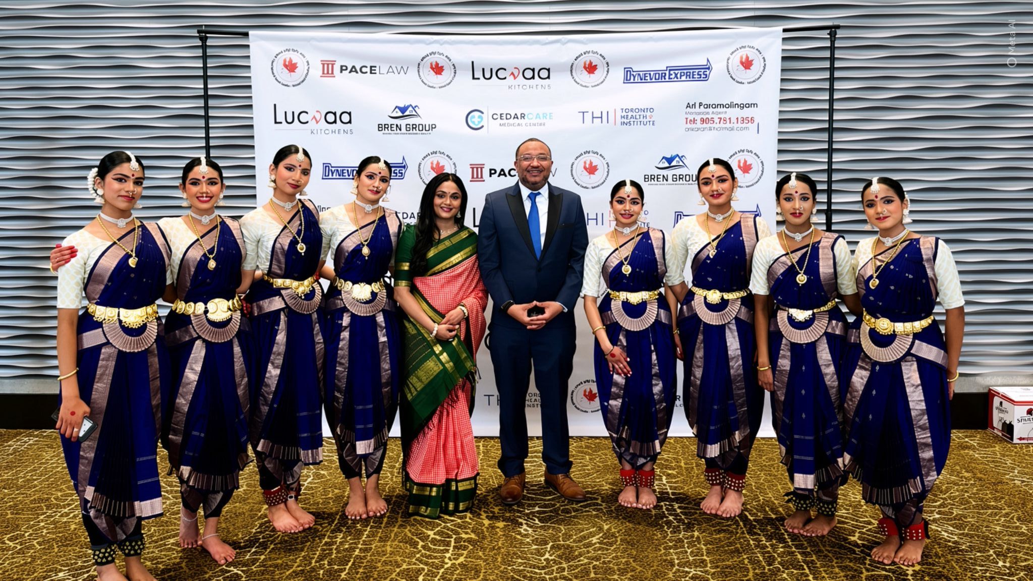 Group of people in traditional clothing standing indoors in front of a screen, posing for a photo.