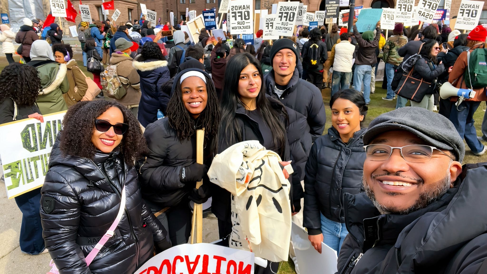 Group of people gathered outdoors at a protest or rally, holding signs and banners, some smiling and talking.