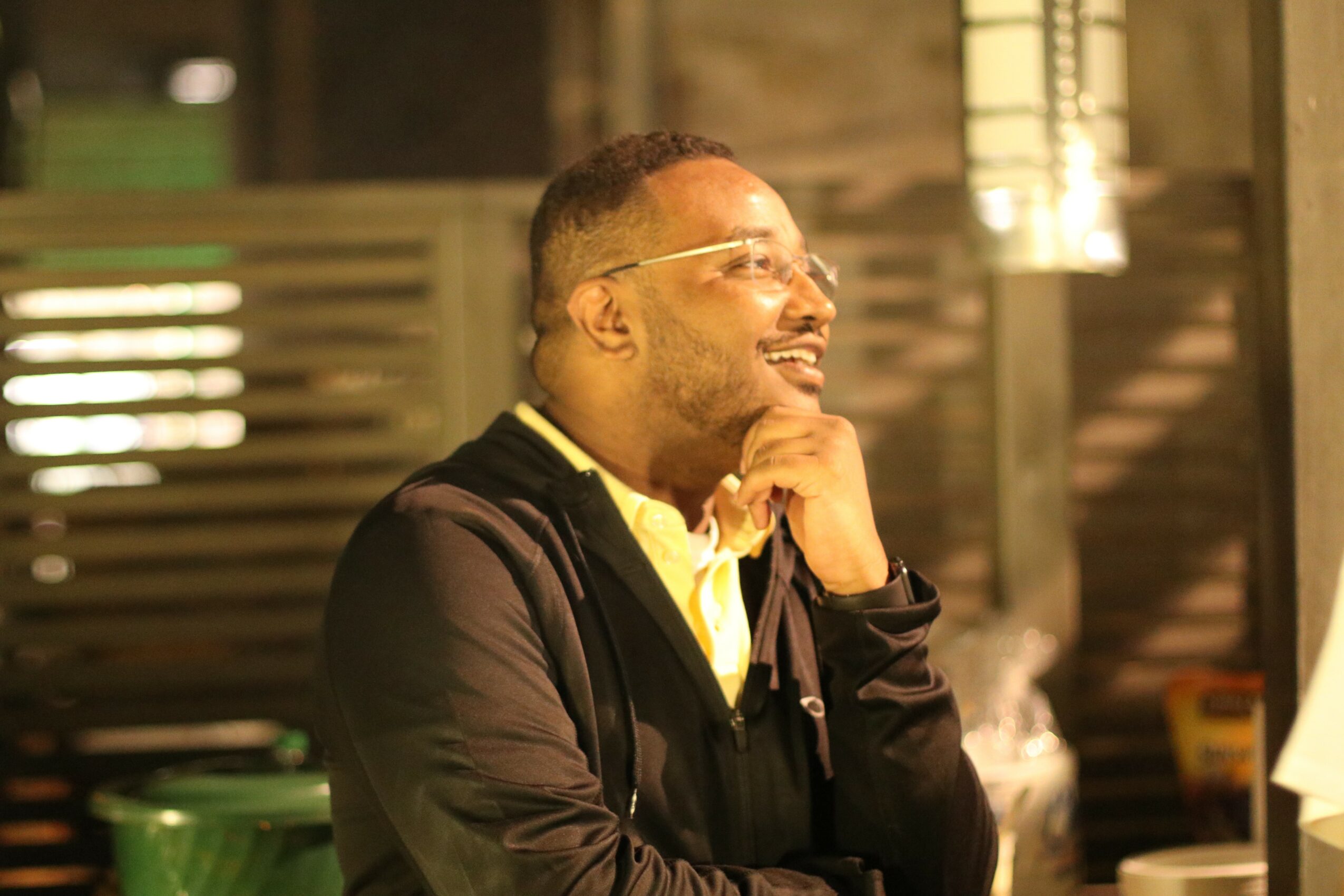 Man smiling with glasses, wearing a black jacket and yellow shirt, indoors with wooden background.
