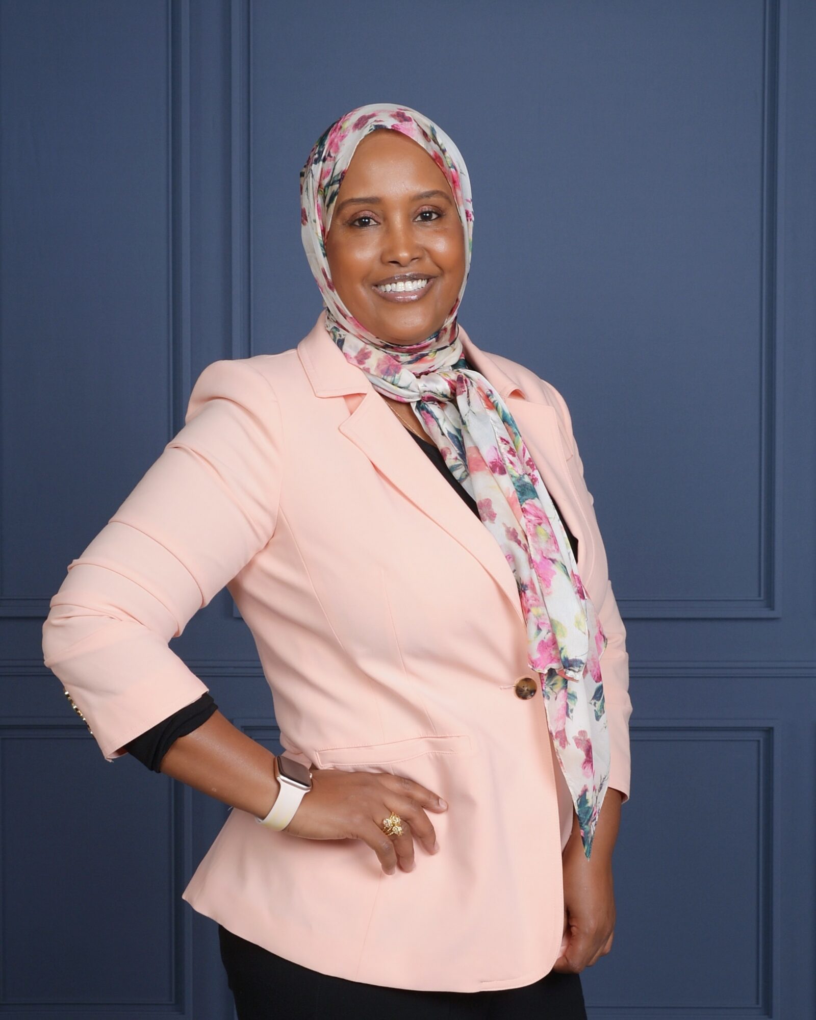 Woman in pink blazer and floral headscarf smiling, standing against a blue wall.