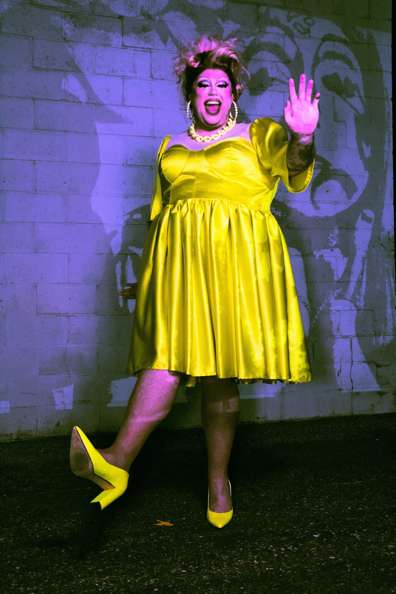 Performer in a bright yellow dress and high heels, gesturing with one hand, standing against a brick wall with shadow patterns.