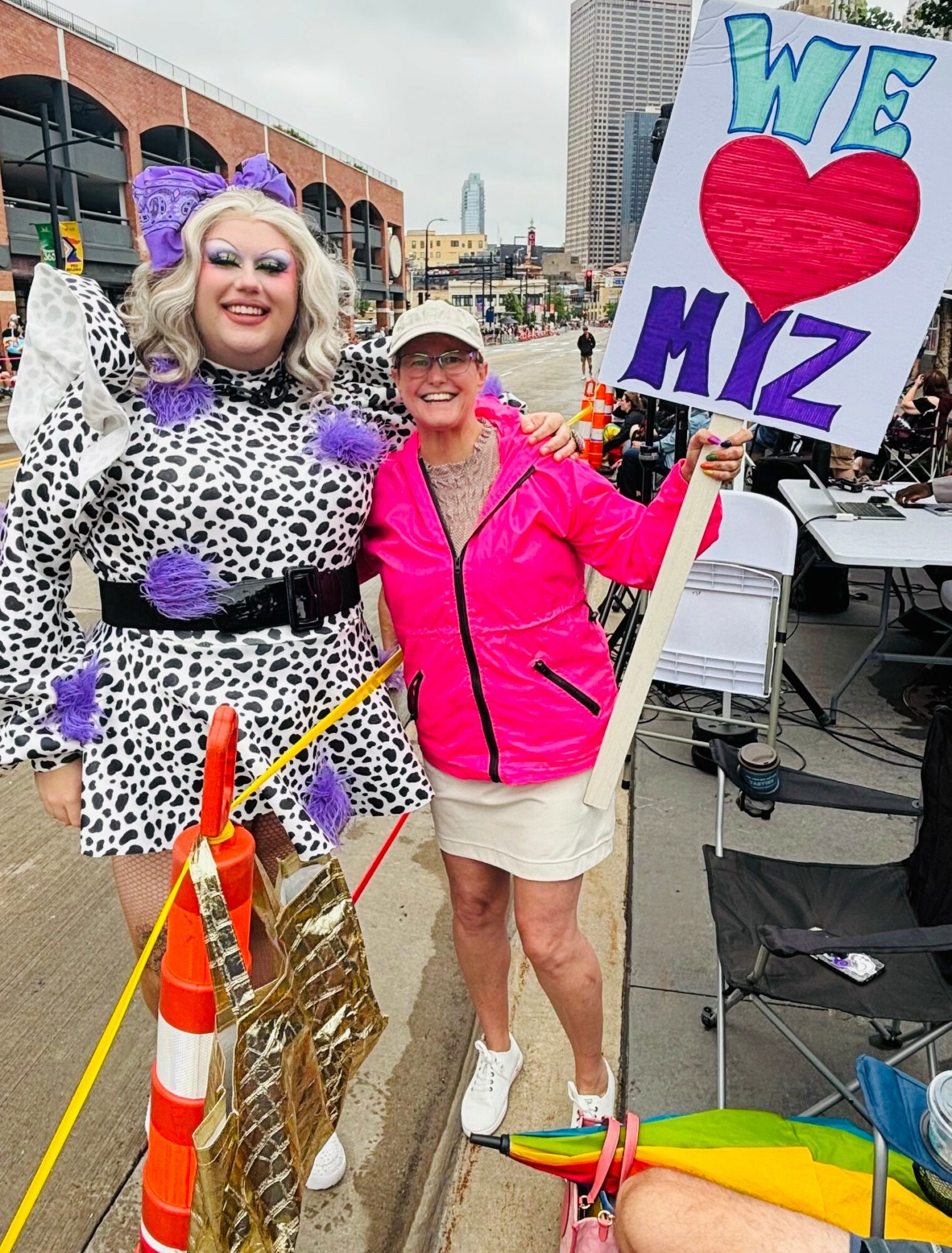Two women smiling outdoors, one in a leopard print outfit with purple accessories, the other in a pink jacket holding a sign that says 