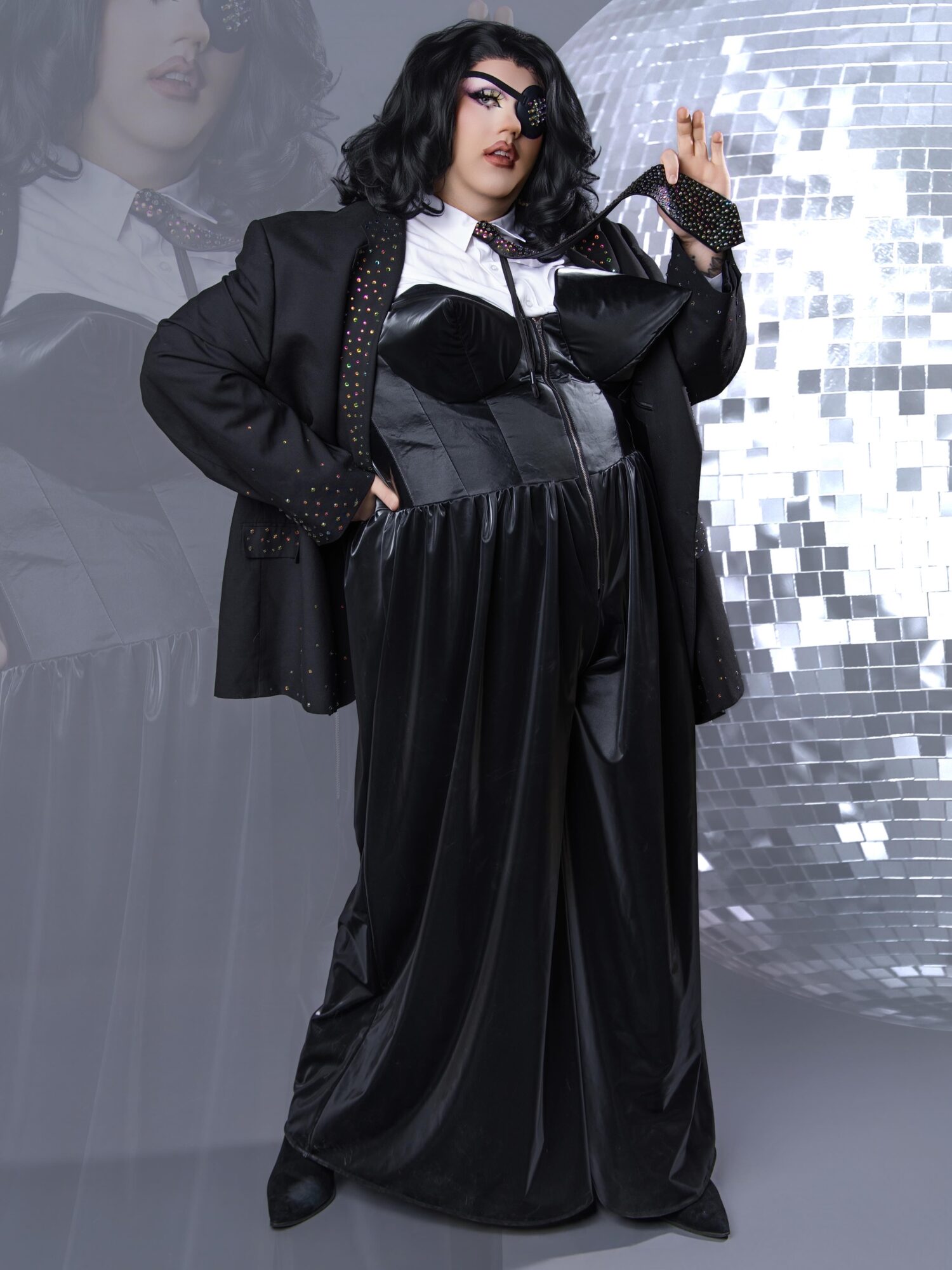 Woman with black hair and glasses in a black outfit, standing in front of a reflective disco ball background.