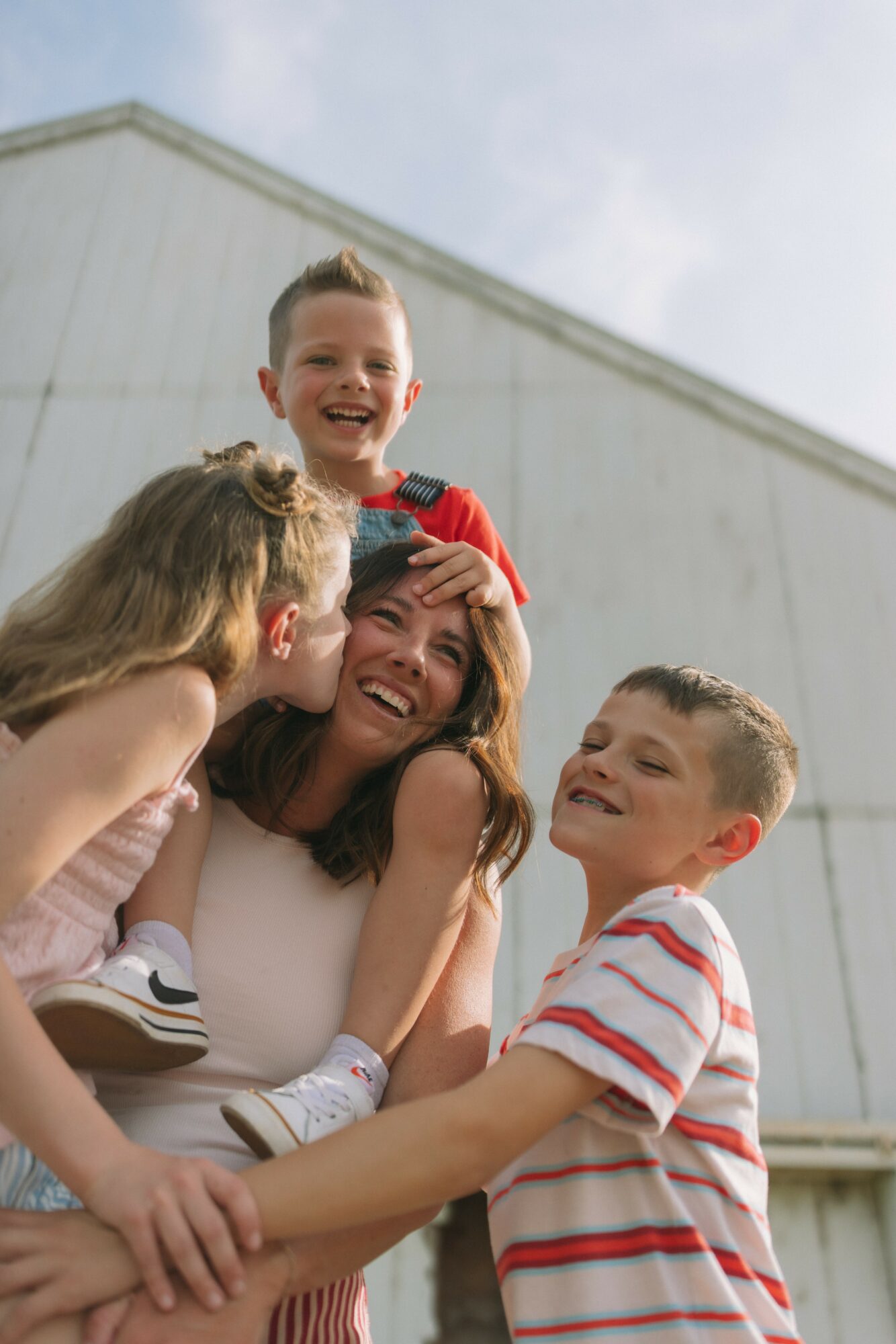 A woman with three children outdoors, all smiling and hugging each other, against a building and sky background.