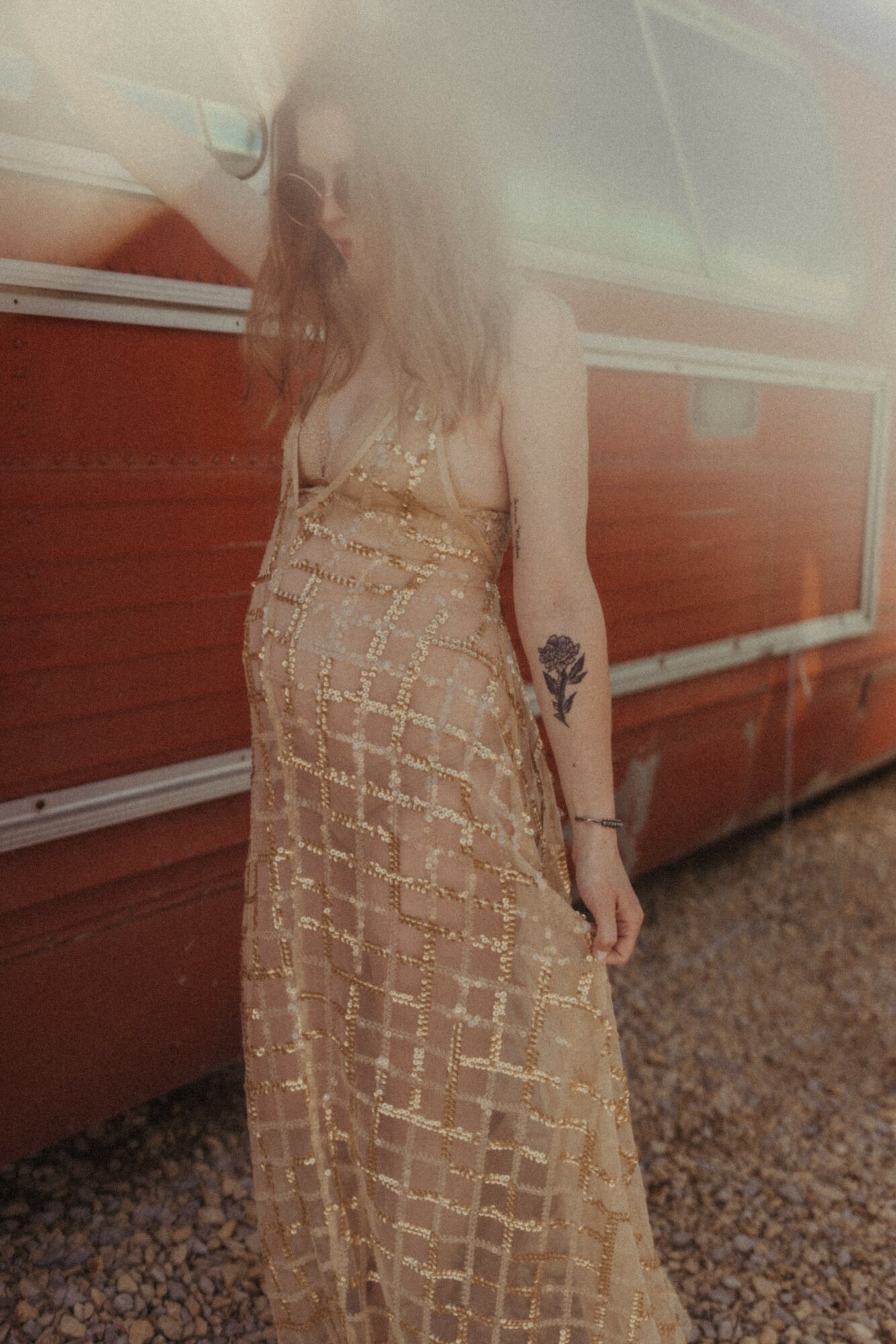 Person with long hair wearing a beige dress with a grid pattern, standing outdoors near a red vehicle.